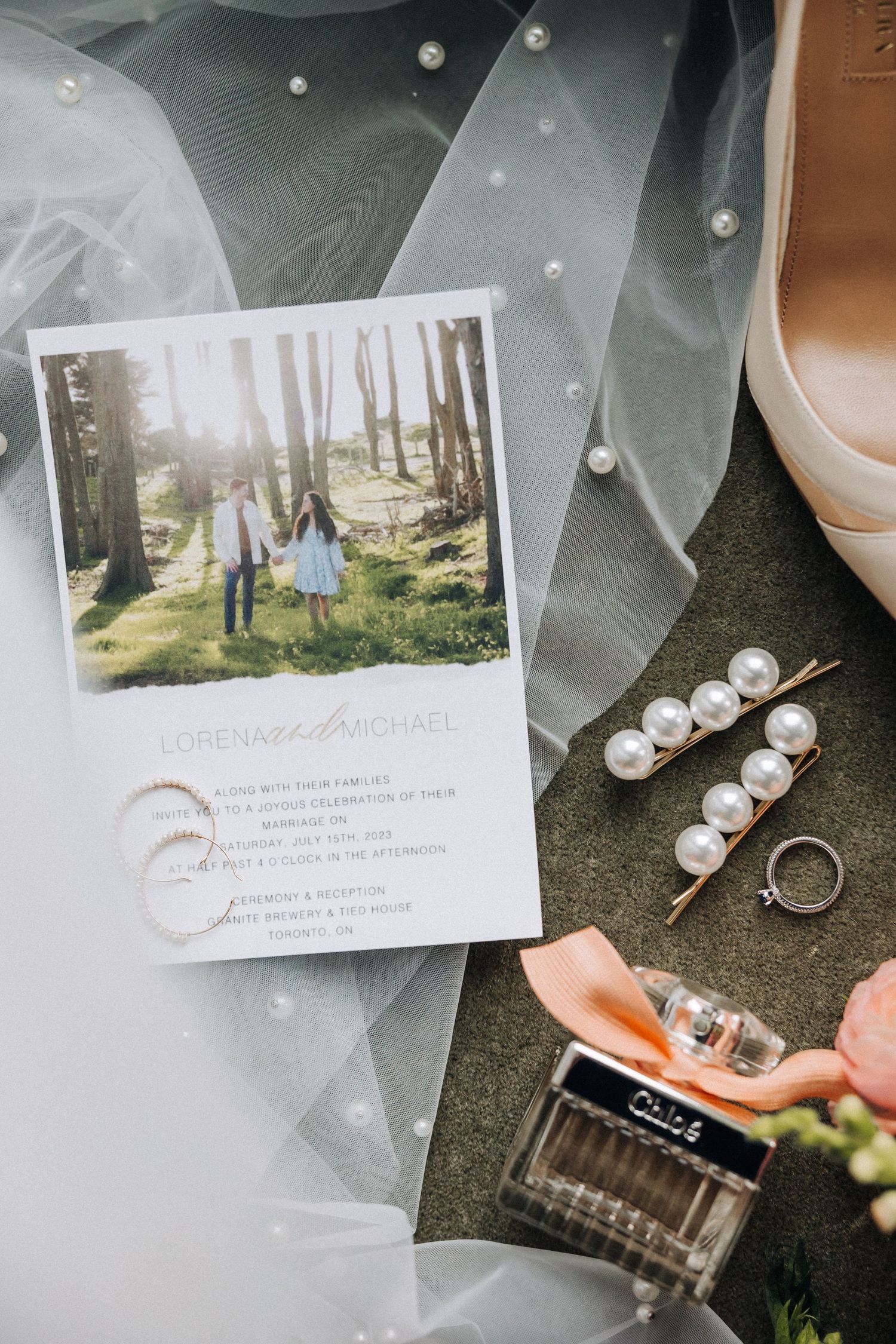 Wedding details including a photo card, pearl hairpins, veil tulle, and perfume bottle arranged on a dark surface.
