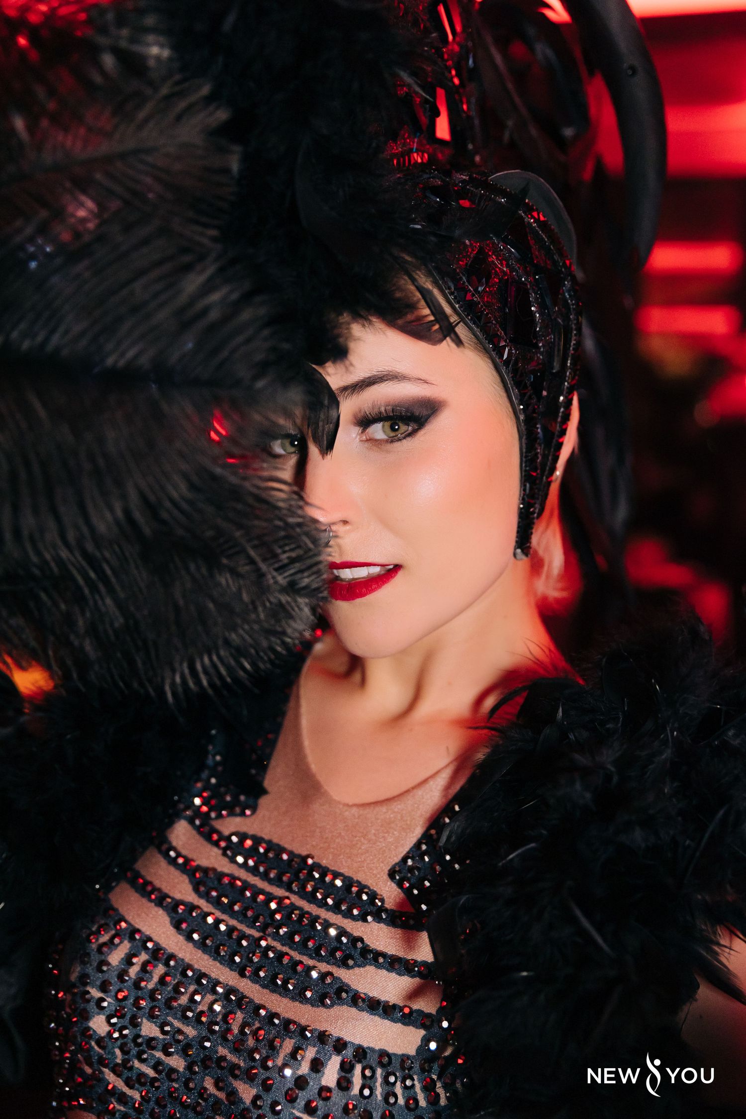 A glamorous cabaret-style performer in black feathers and sequins poses against a red-lit backdrop.