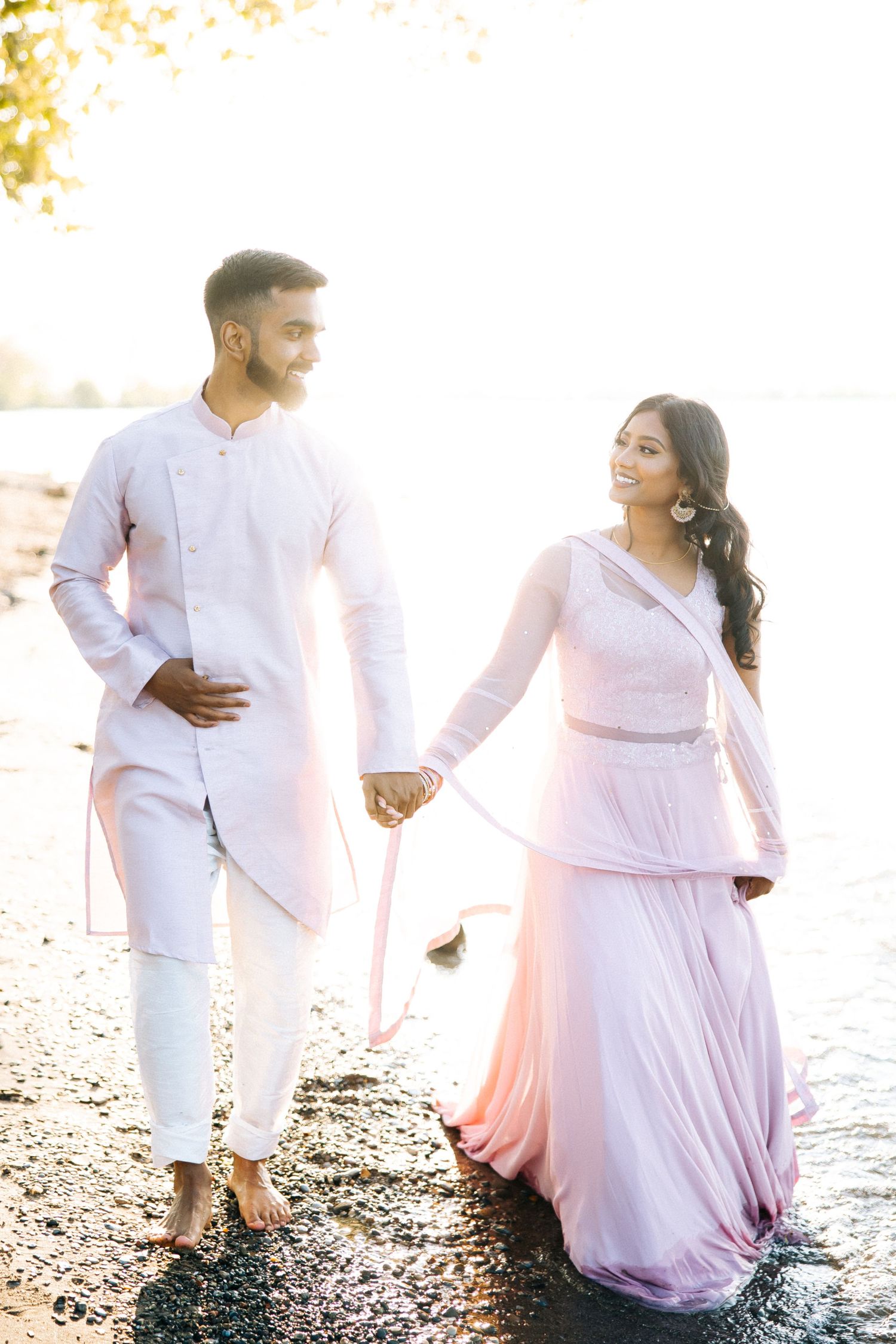 A couple in coordinated white and pink outfits walks along the waterfront at sunset.