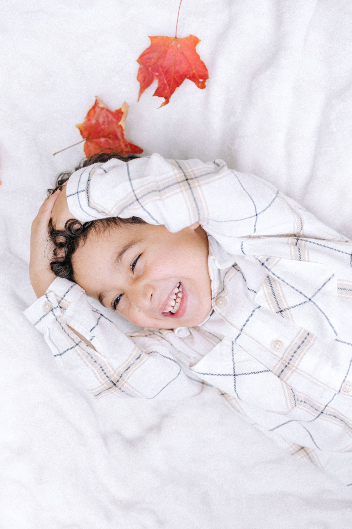 A child lies on white bedding with red maple leaves overhead, smiling brightly.