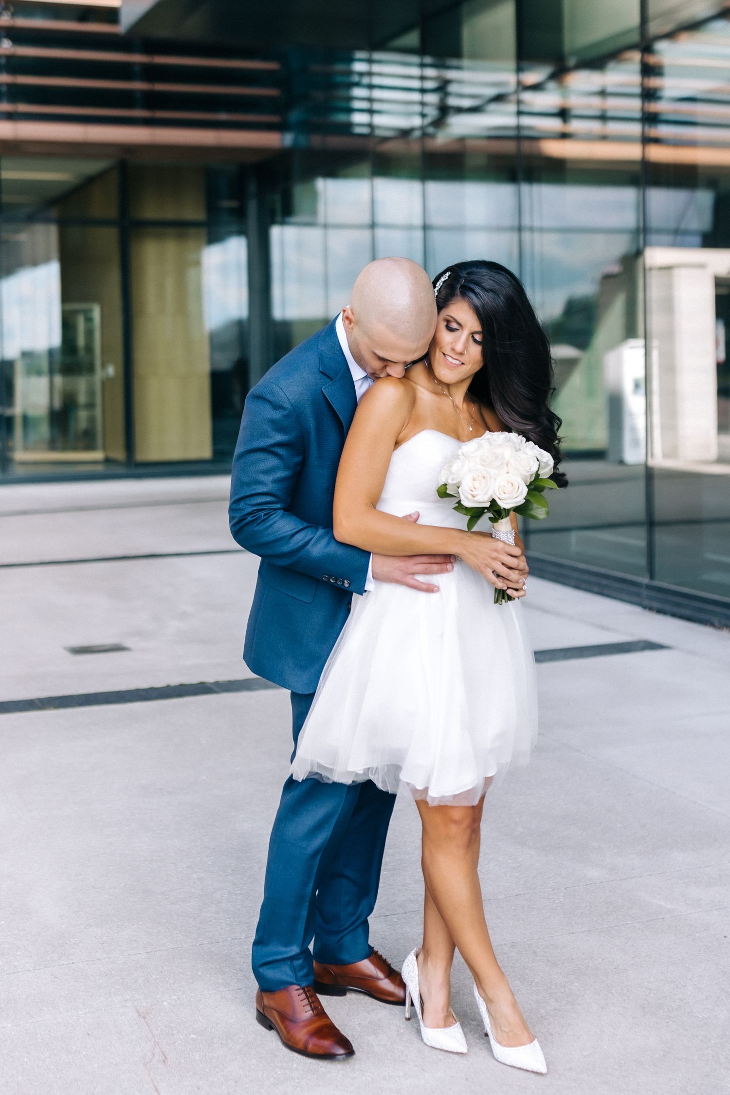 A couple embraces outdoors in wedding attire - she in a short white dress holding white flowers, he in a blue suit.