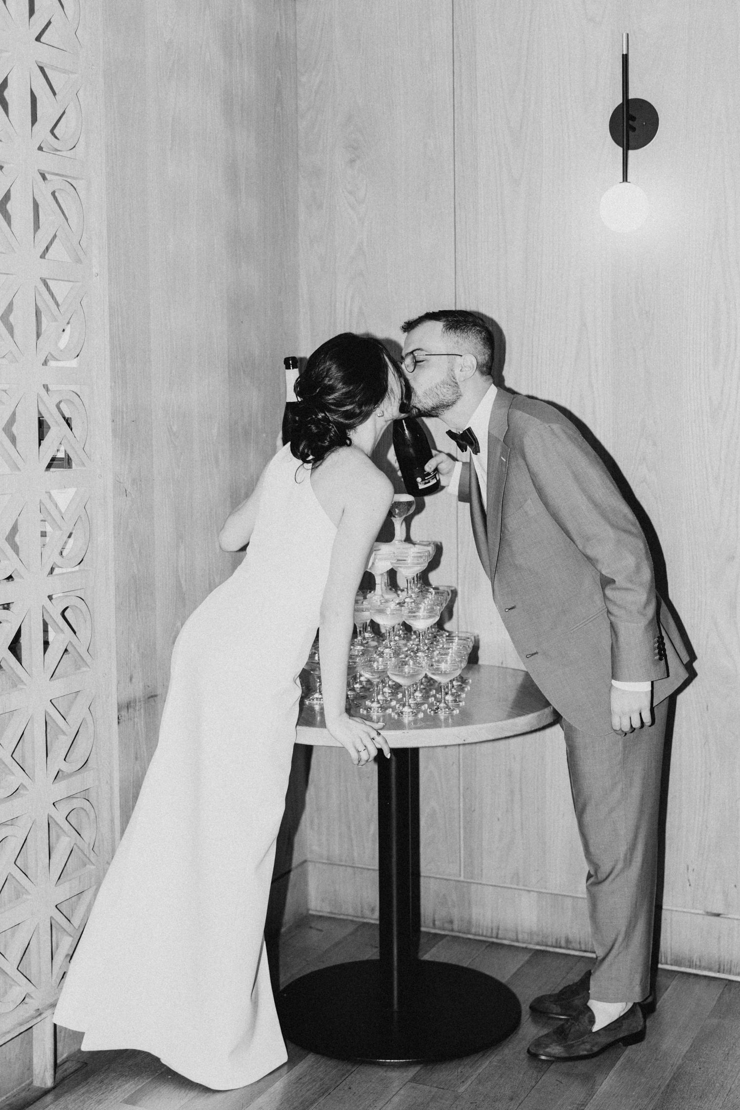 A black and white vintage wedding photo showing a couple kissing over their champagne tower at their reception.
