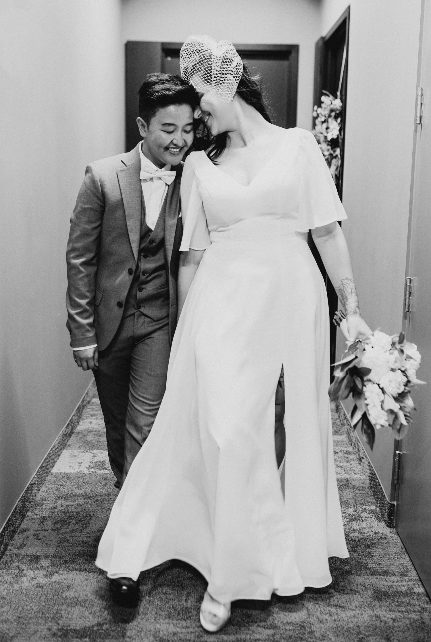 Black and white wedding photo showing couple walking down a hallway, with one in a suit and the other in a flowing jumpsuit.