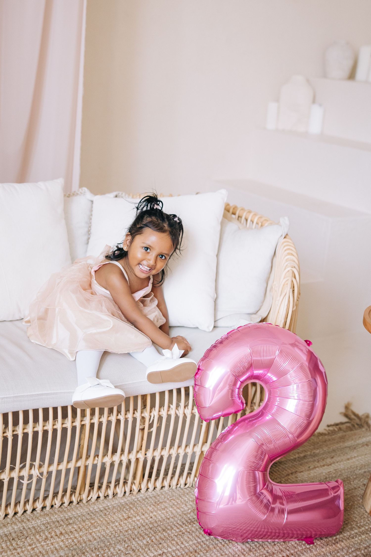 A young child in a tulle dress poses near a pink number 2 balloon on a couch.