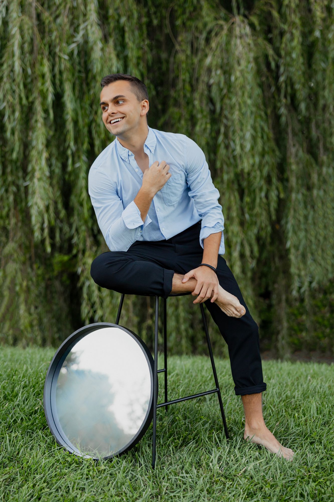 Casual outdoor portrait with person in rolled-up sleeves sitting on stool against weeping willow backdrop.
