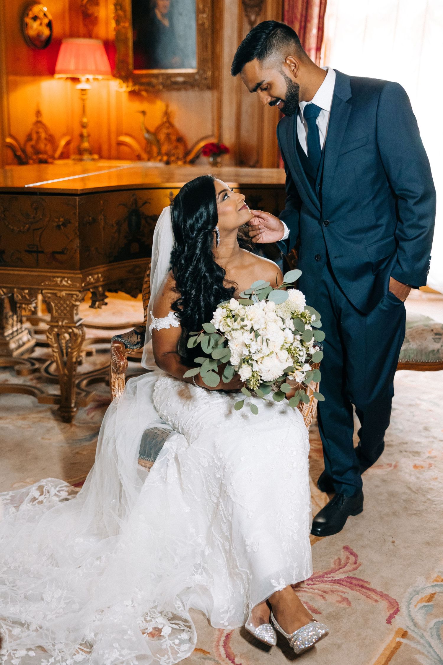 A bride in a white lace gown holds a eucalyptus bouquet while sharing an intimate moment in an ornate room with warm lighting.