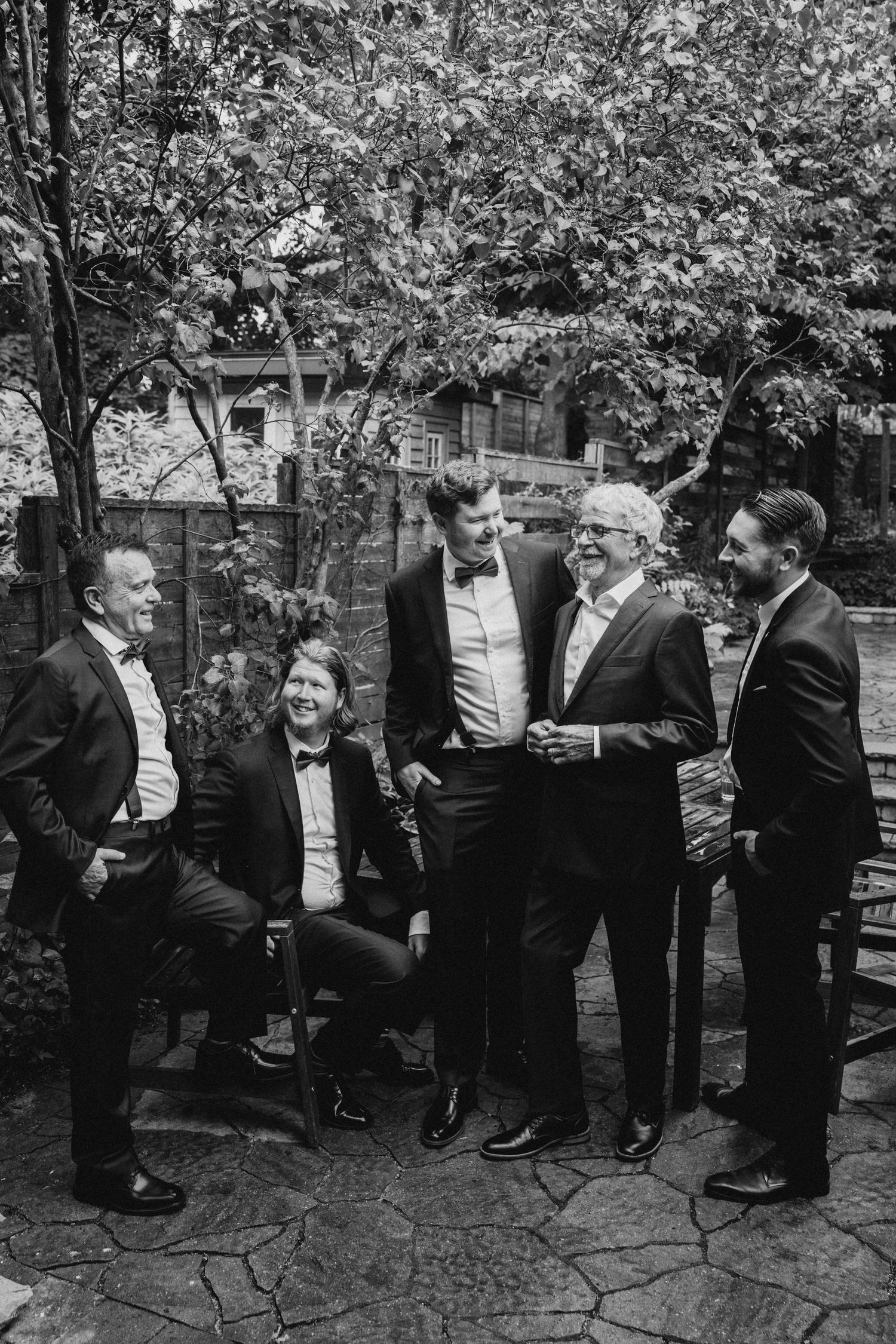 A black and white photo of a group of men in formal attire laughing together outdoors on a stone patio.