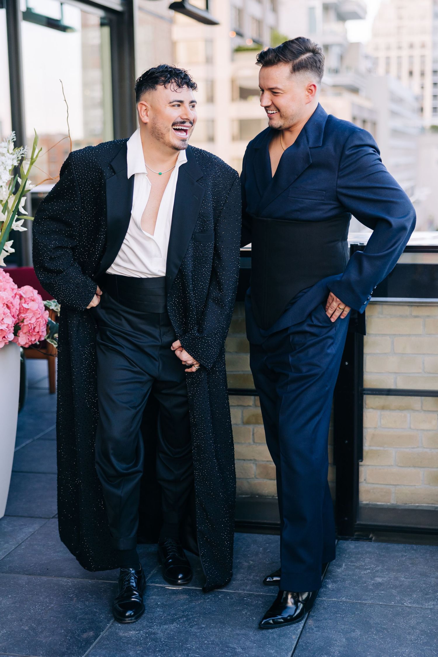 Two people in elegant navy suits share a joyful laugh while standing outdoors on a terrace with pink flowers.