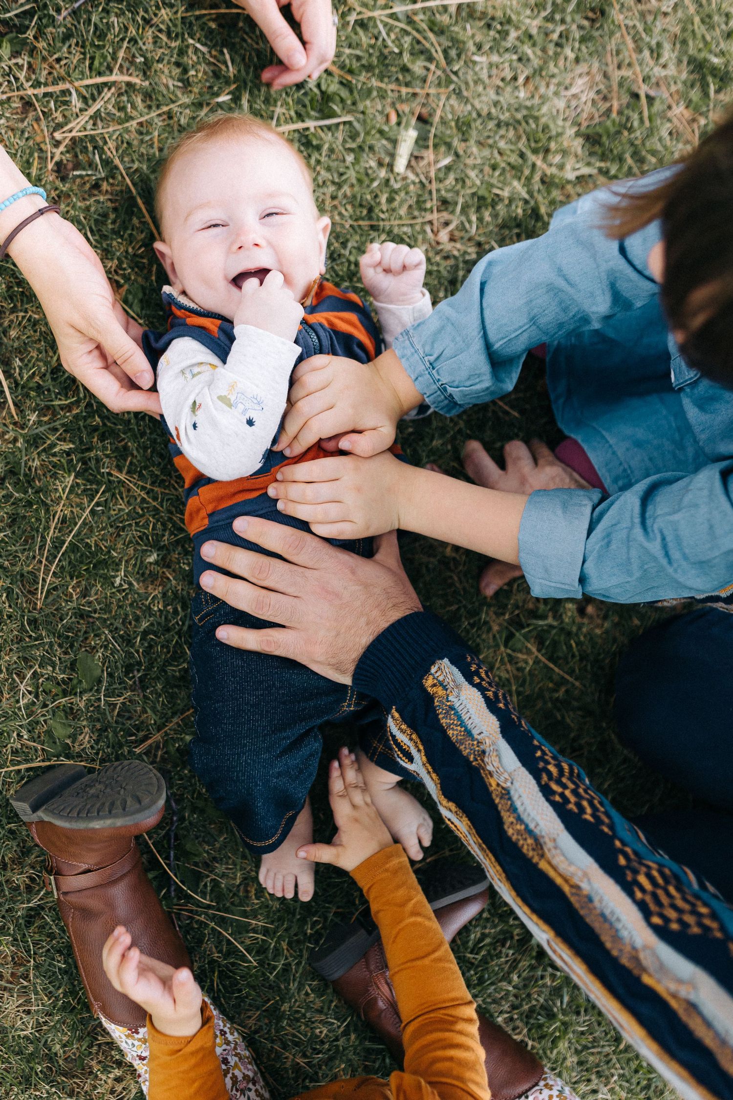 Happy baby lying on grass being tickled by multiple hands.