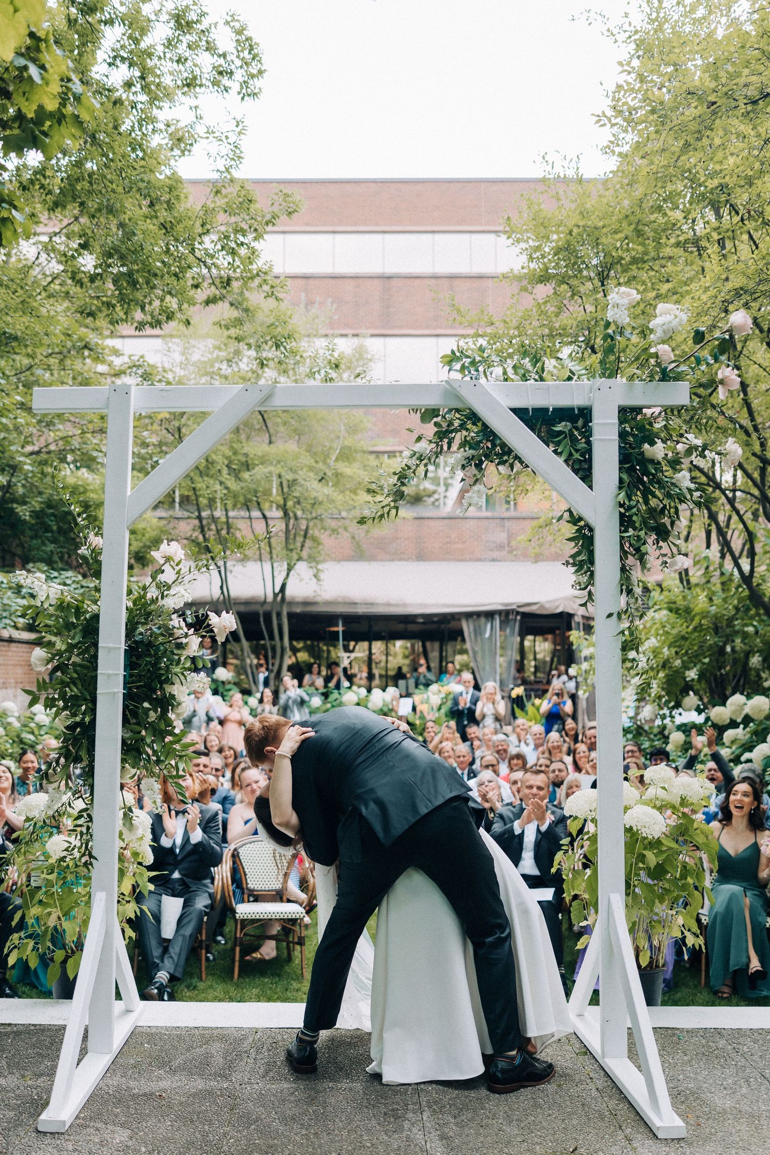 Wedding couple shares a romantic dip kiss under a white arch as guests watch in an outdoor garden ceremony.