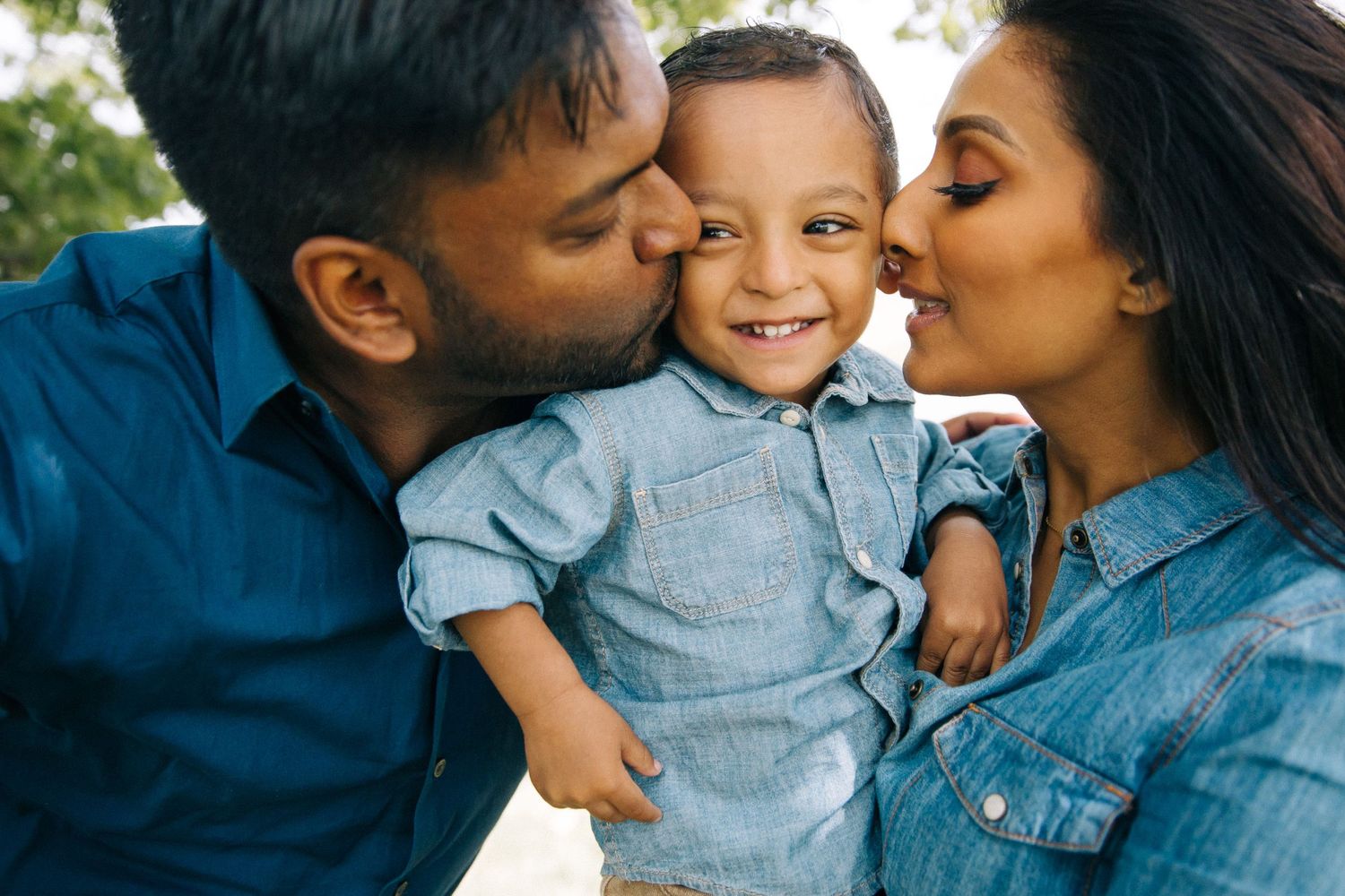 Parents giving their young child loving kisses on both cheeks outdoors.