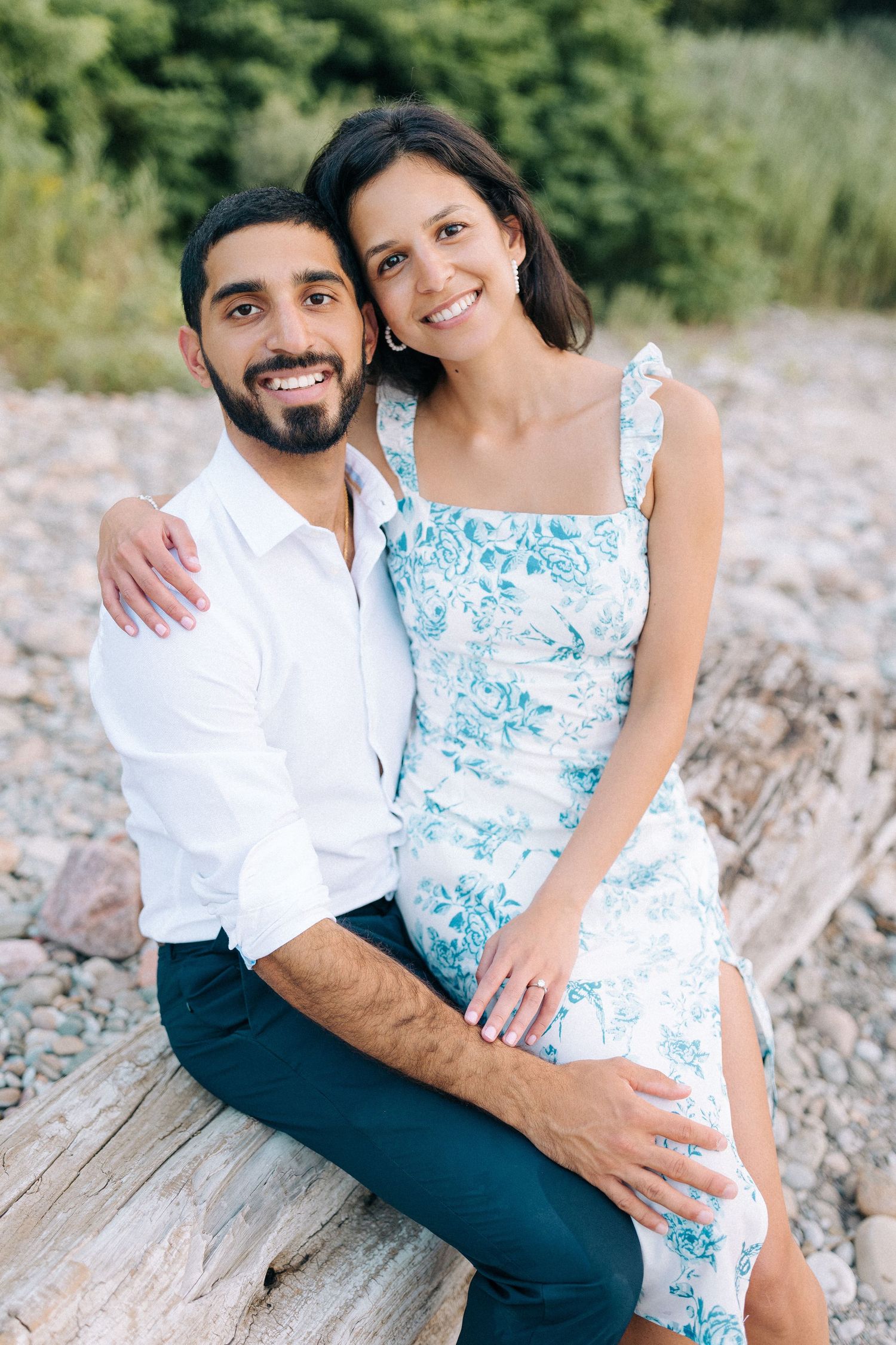 A couple sits together outdoors on driftwood, wearing a white shirt and blue floral summer dress against a natural backdrop.