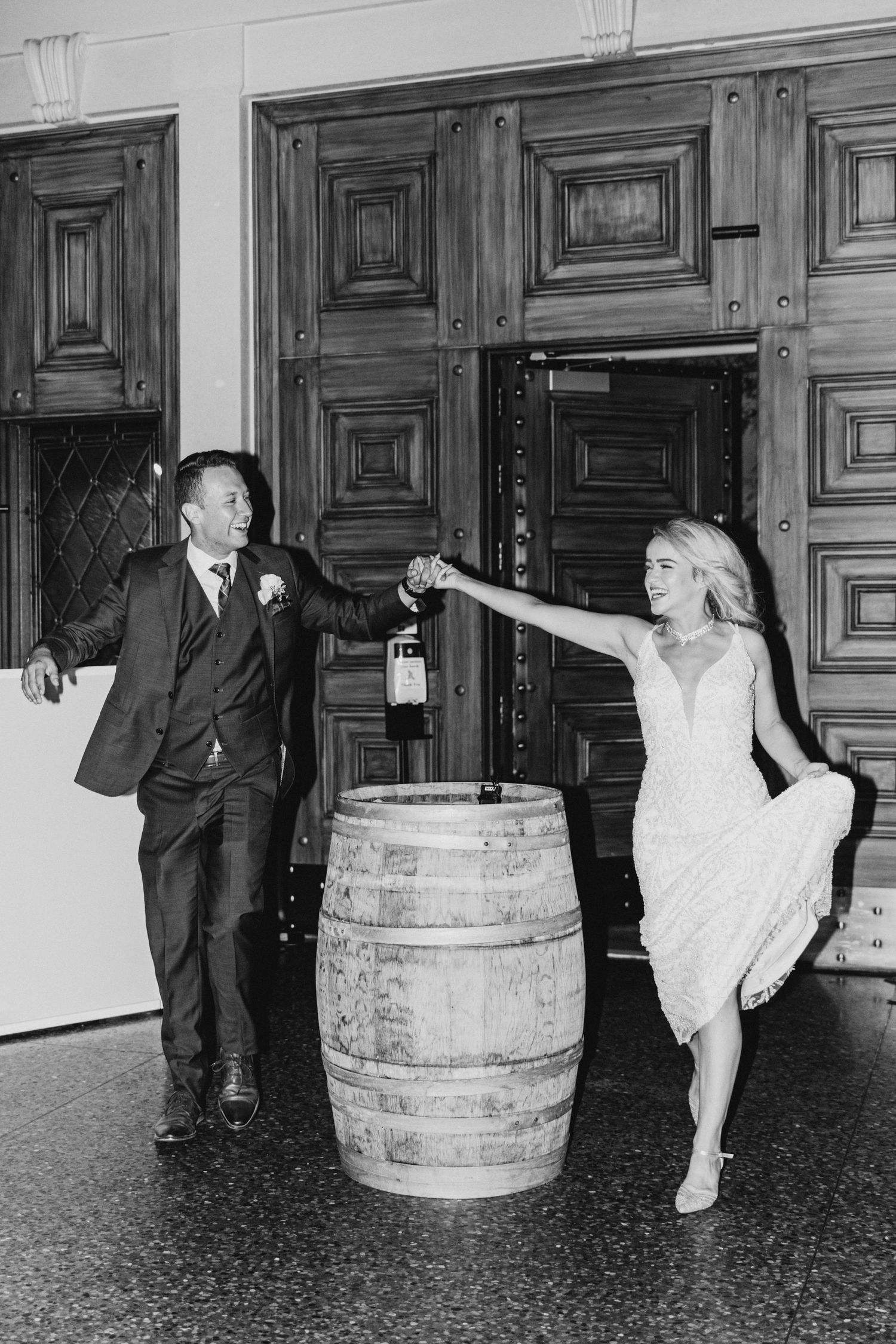 A couple playfully poses with a wine barrel at a rustic wedding venue, with the bride wearing a white dress.