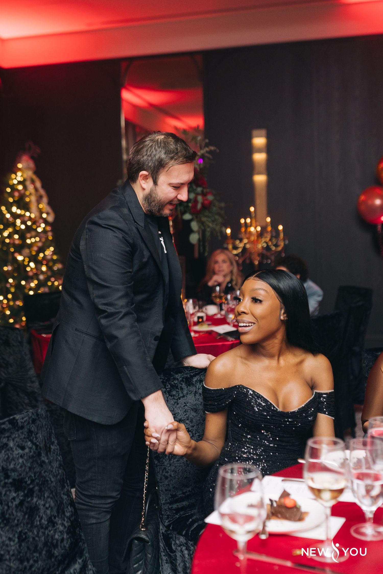 Two people share an intimate moment at an elegant holiday dinner party with Christmas decor and red table settings.