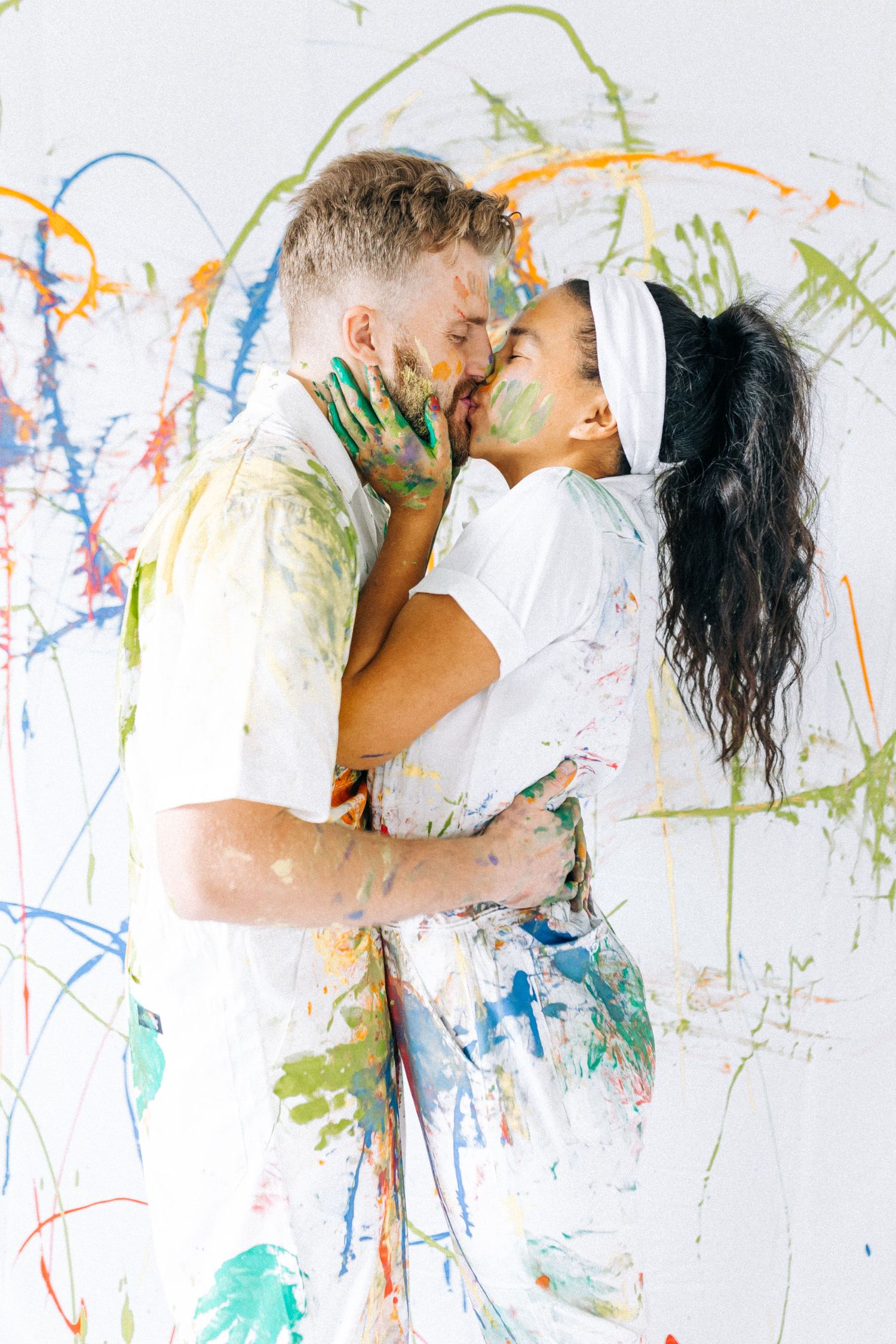 Couple covered in colorful paint embracing against splatter-painted wall.