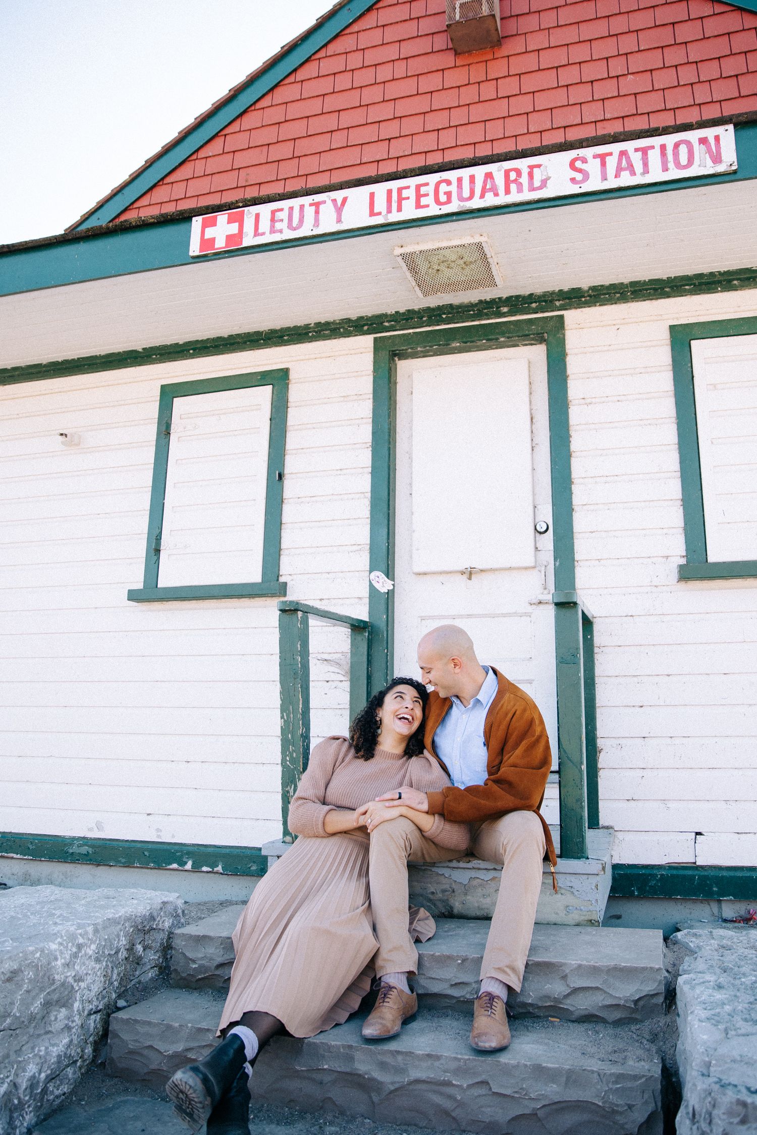 A couple sits together on the steps of a white lifeguard station building.