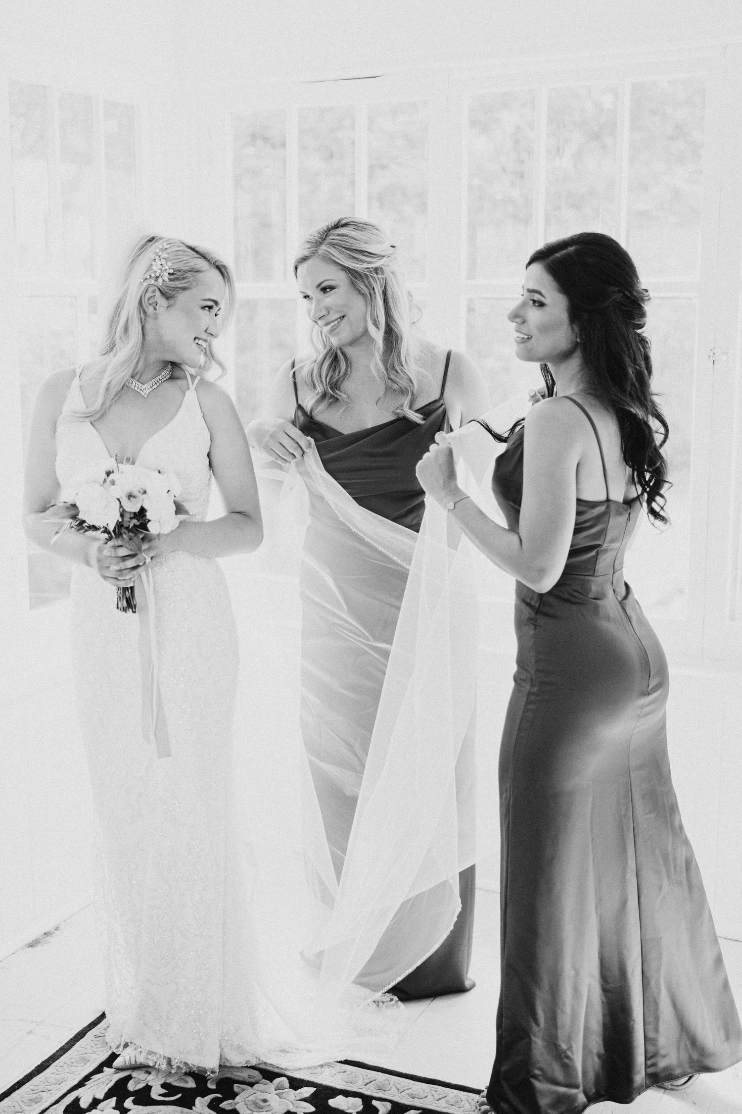 Black and white photograph of three women in elegant dresses, with one in bridal white holding a bouquet.