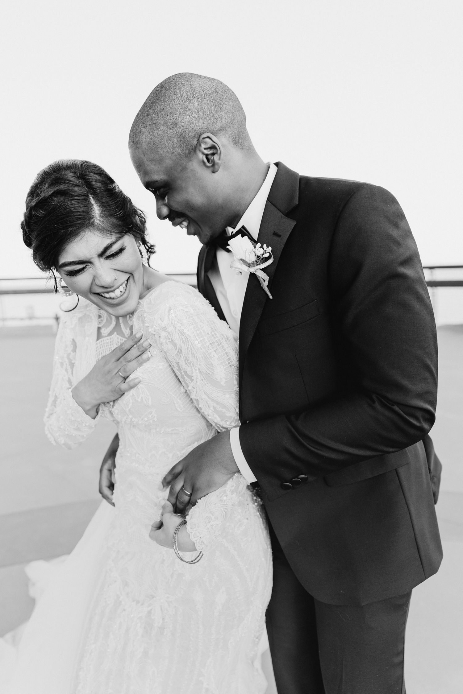 A joyful black and white wedding photo capturing an intimate candid moment of laughter between newlyweds outdoors.