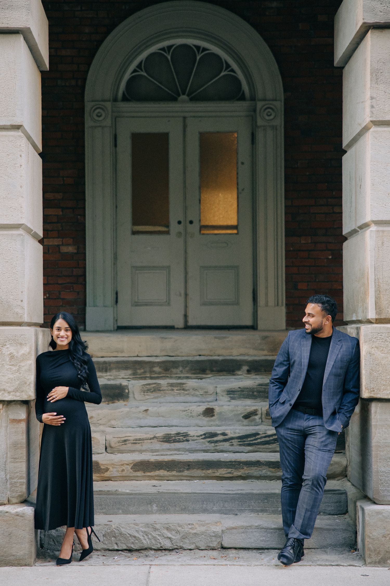A couple stands on stone steps in front of a large arched doorway, with the woman wearing a long black dress.