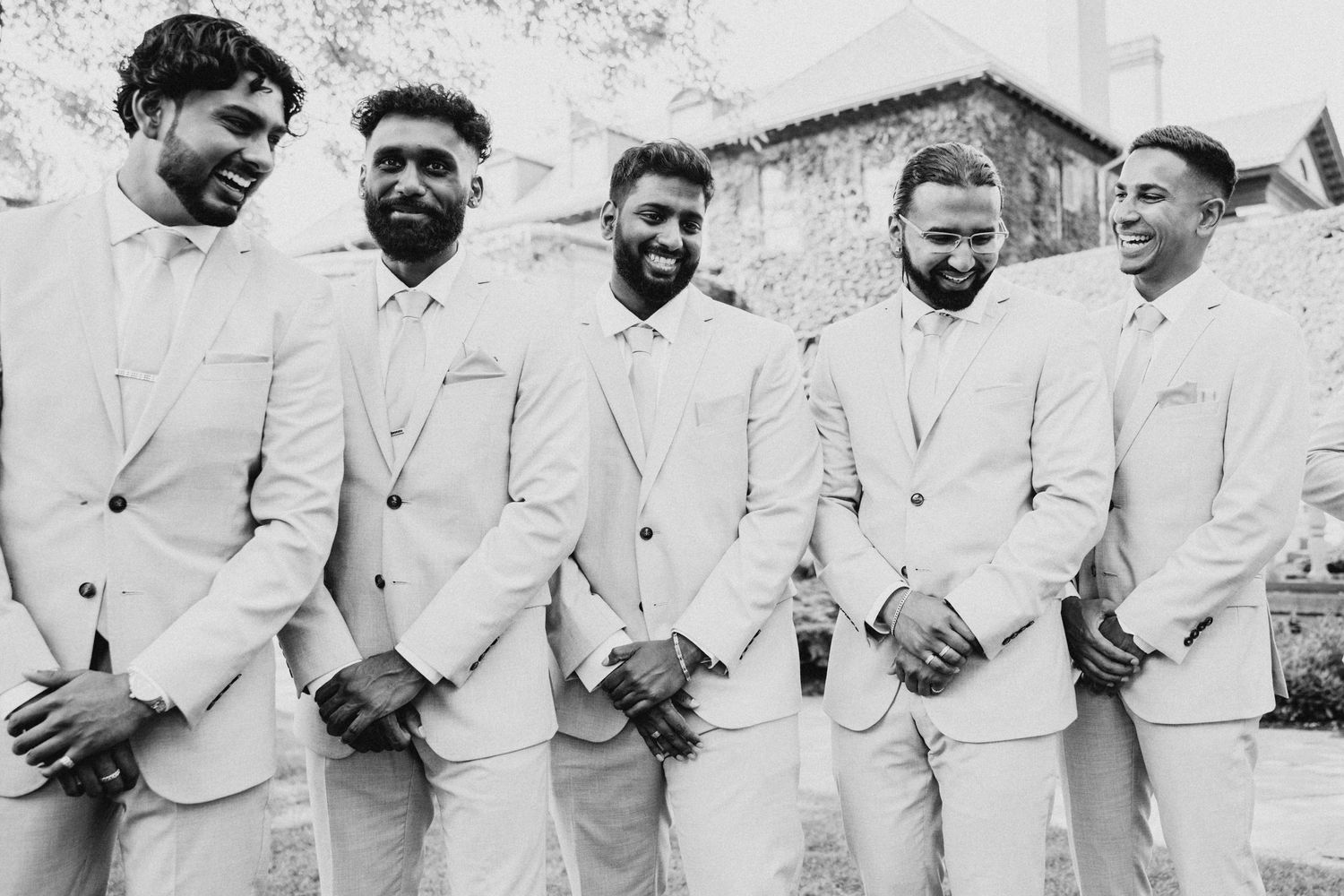 Five people stand in a row wearing matching light-colored suits and smiling in this vintage black and white photograph.