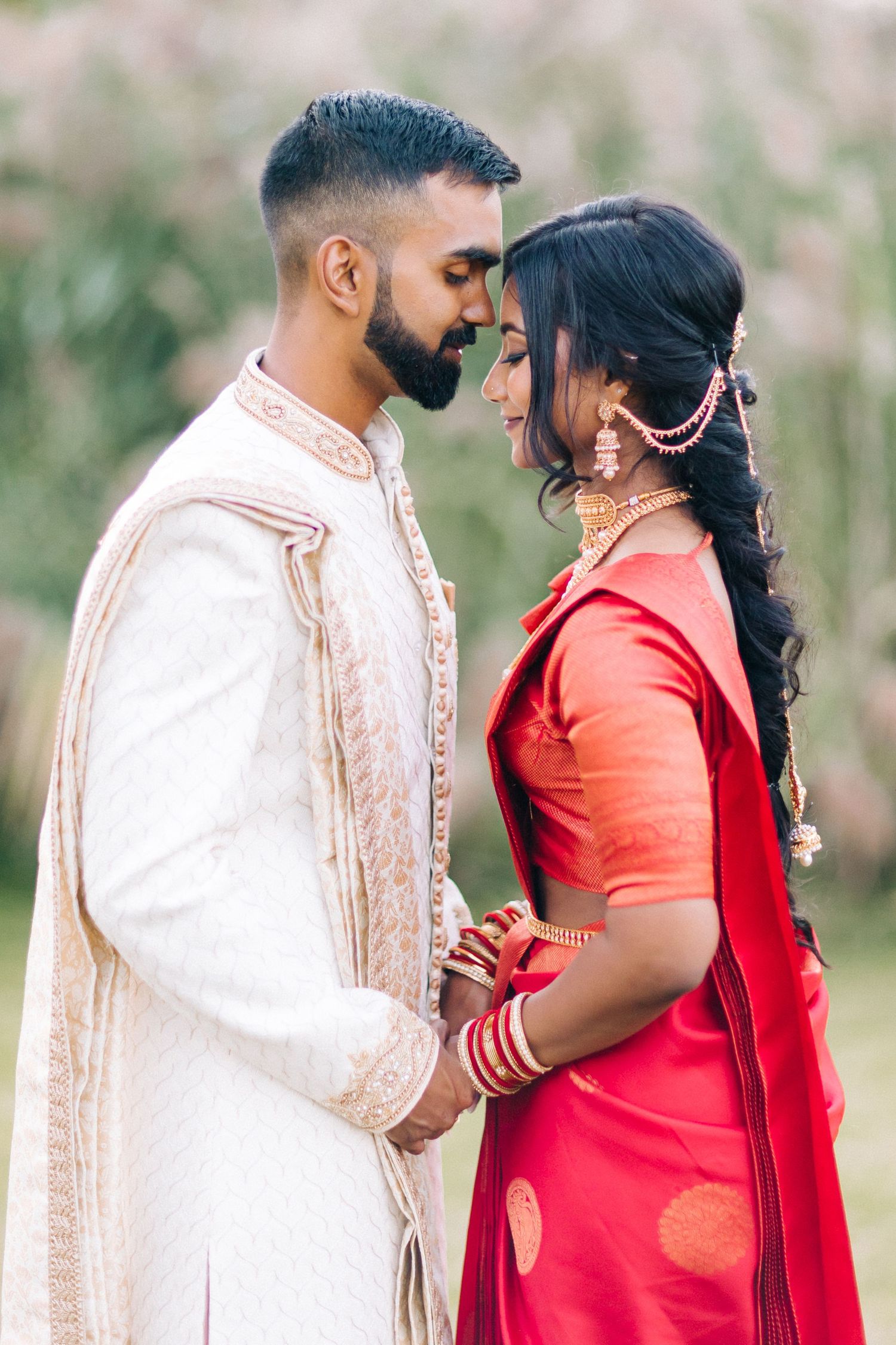 A couple in traditional Indian wedding attire share an intimate moment outdoors, wearing red sari and cream sherwani.