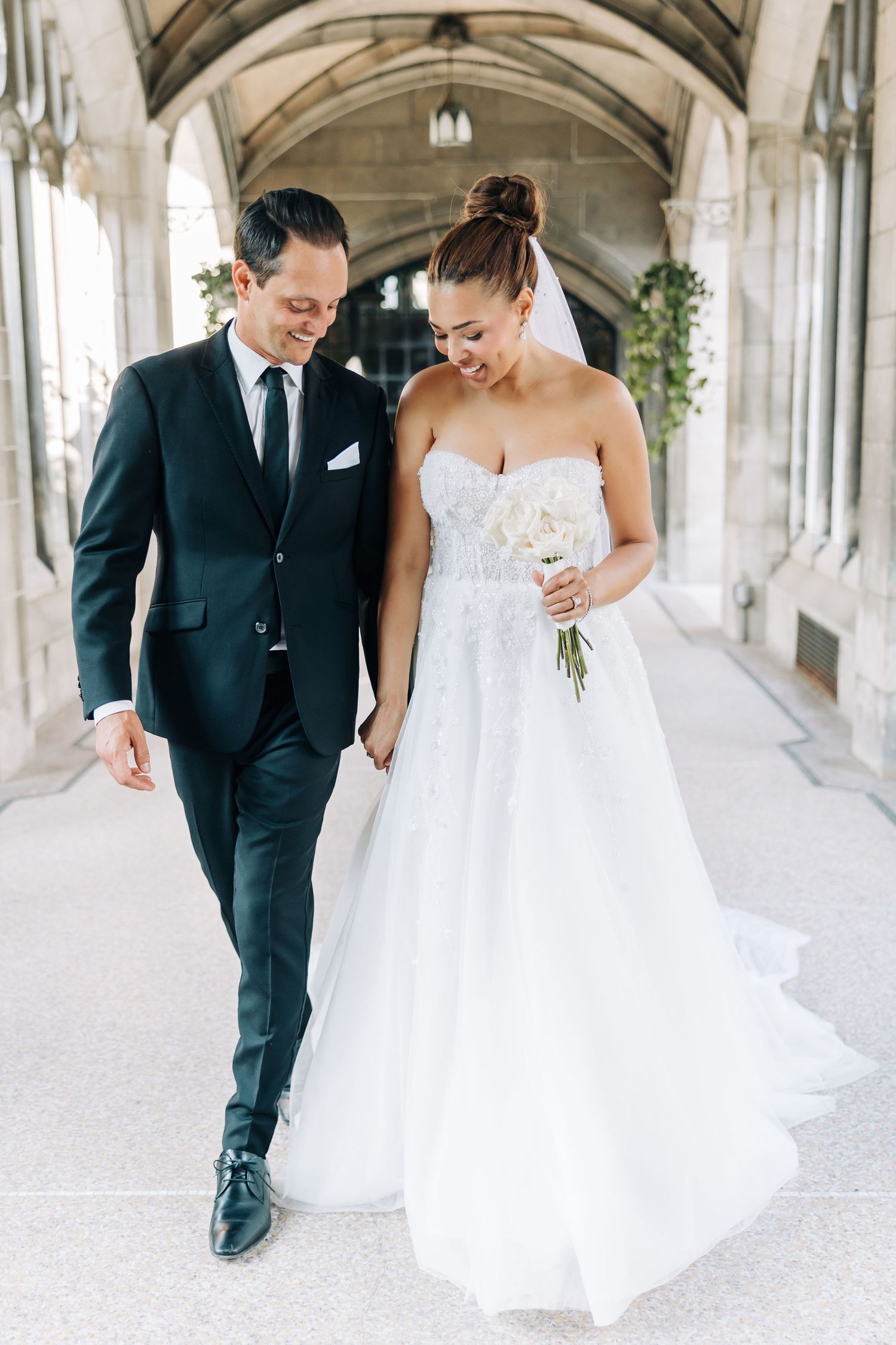 A couple walks together in an elegant stone archway corridor, the bride in a flowing white gown and groom in dark suit.