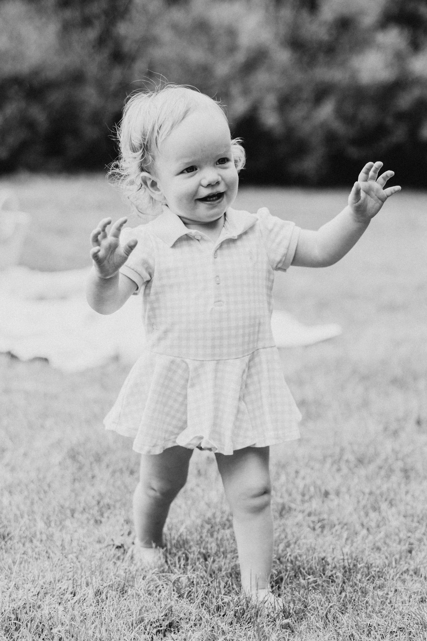 Toddler in checkered dress playing and laughing outdoors in black and white photo.