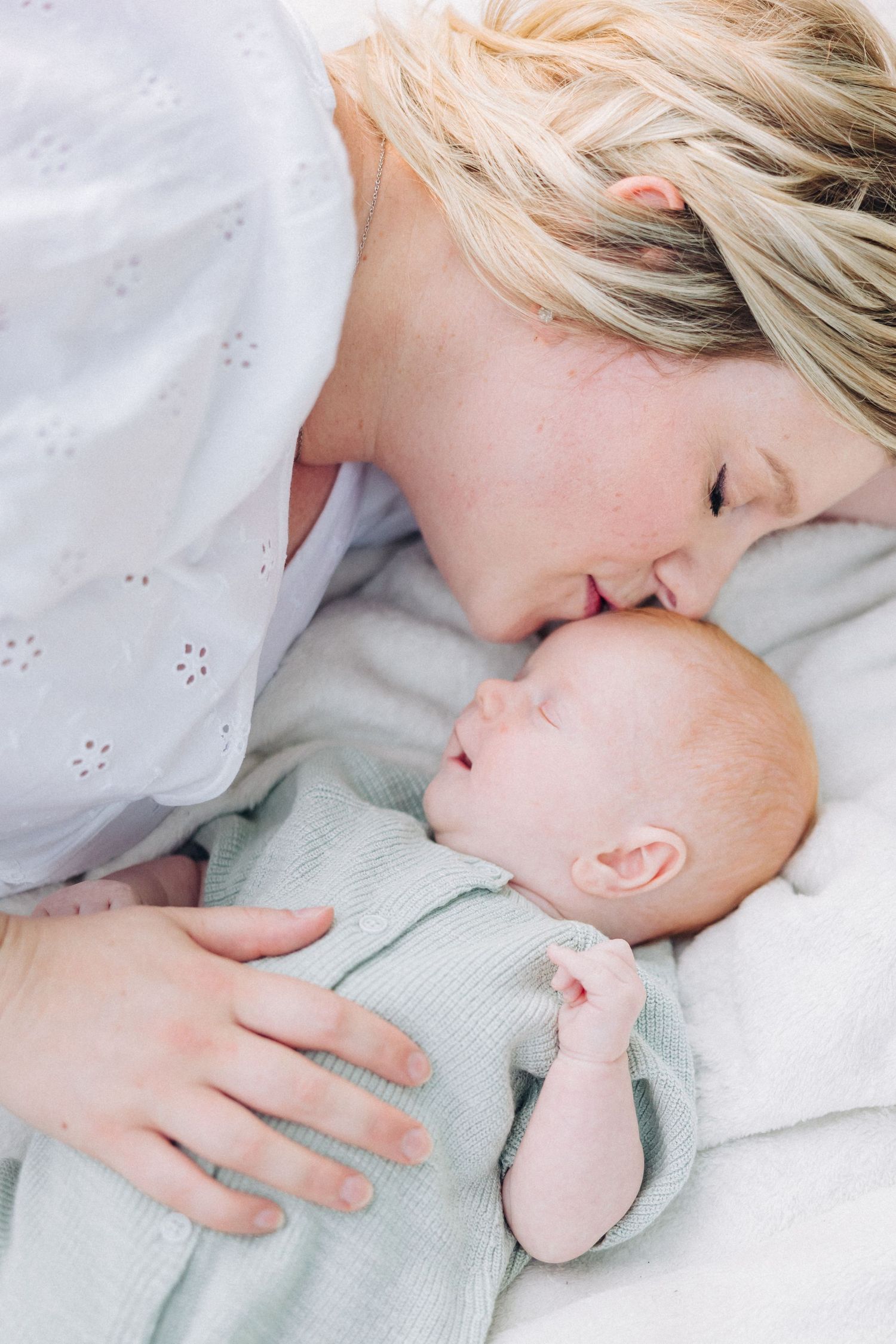 Tender moment between mother and newborn baby lying together on white bedding.