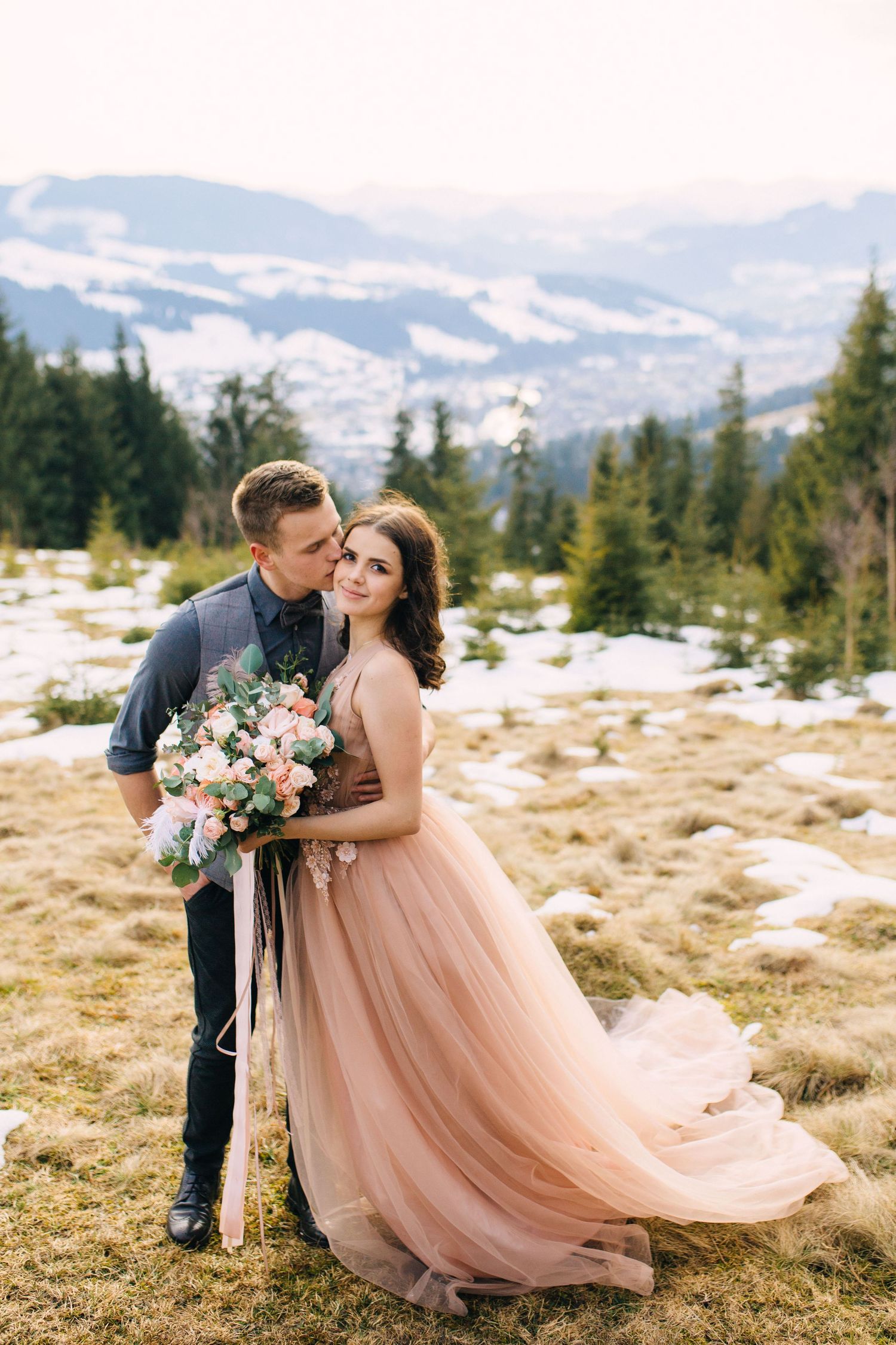 Romantic mountain engagement photo with flowing blush gown and floral bouquet against snowy mountain landscape.