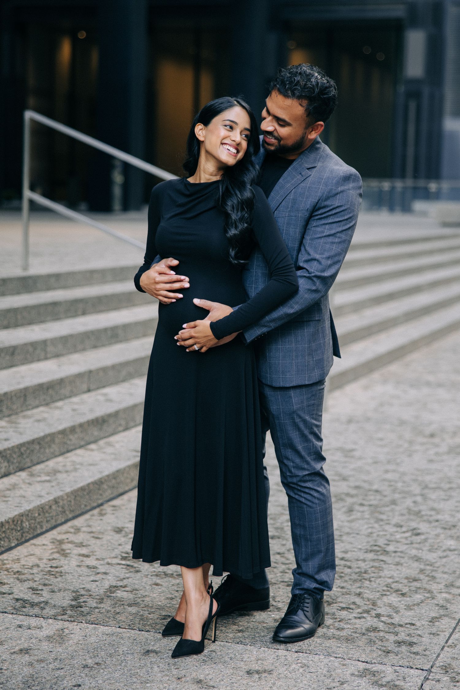 A couple embraces on stone steps, with her wearing a black dress and him in a grey suit.
