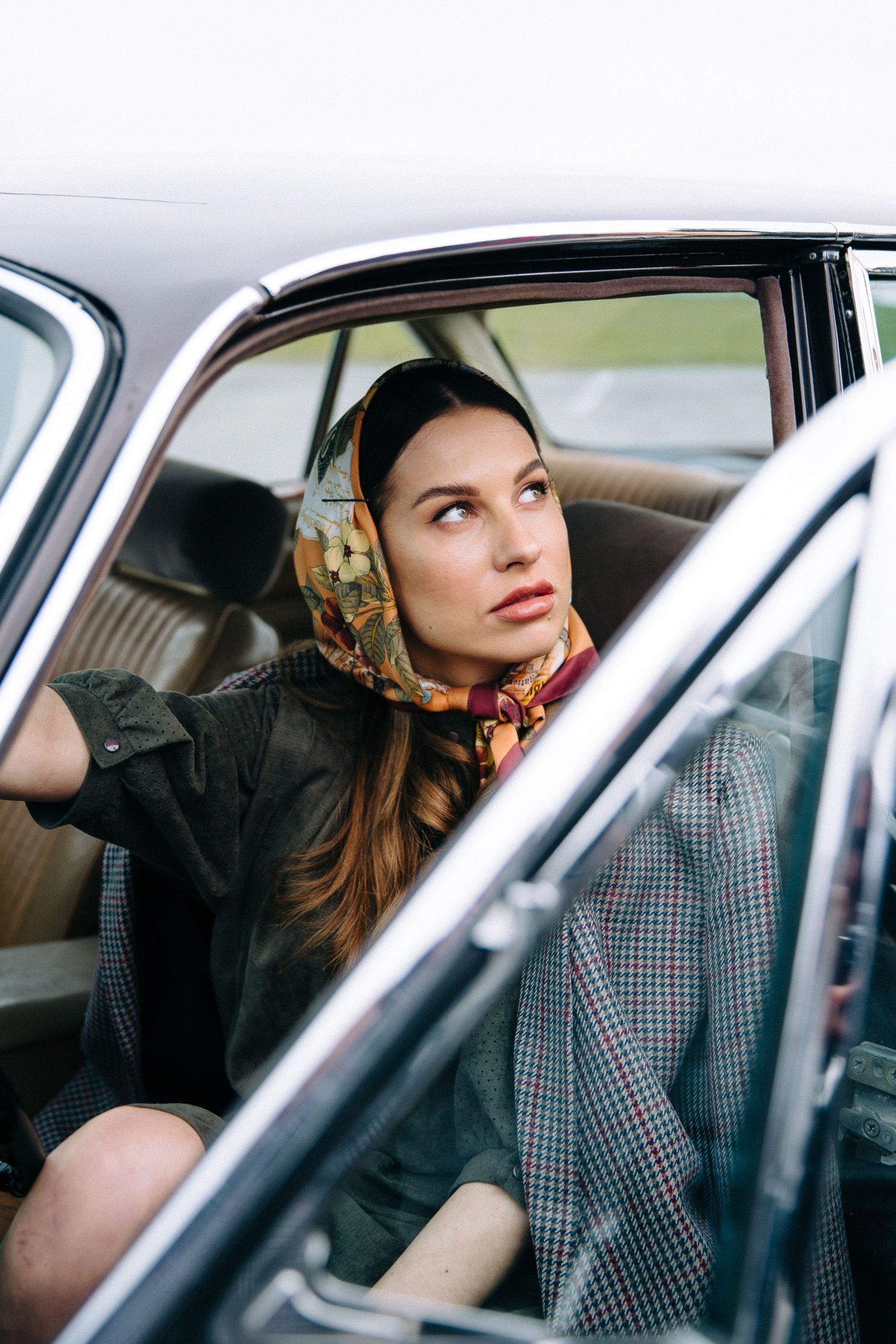 A retro-styled woman in a headscarf sits thoughtfully in a vintage car.