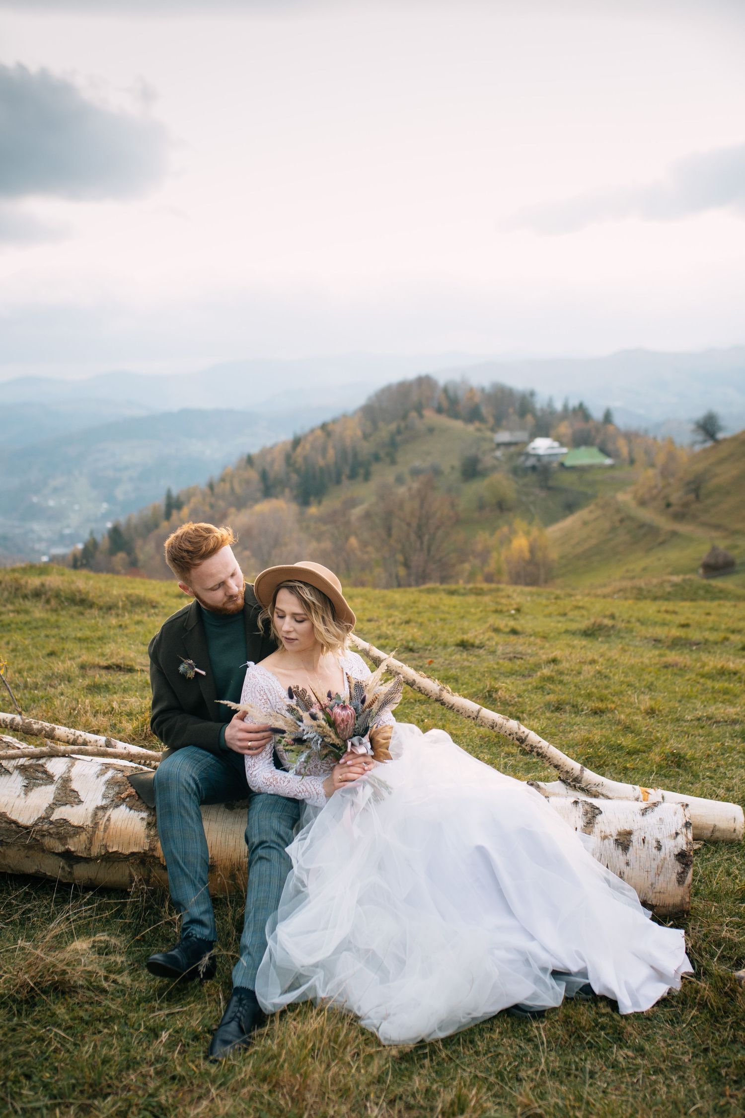 A couple in wedding attire sits on birch logs overlooking mountains on an autumn hillside with a rustic cabin in distance.