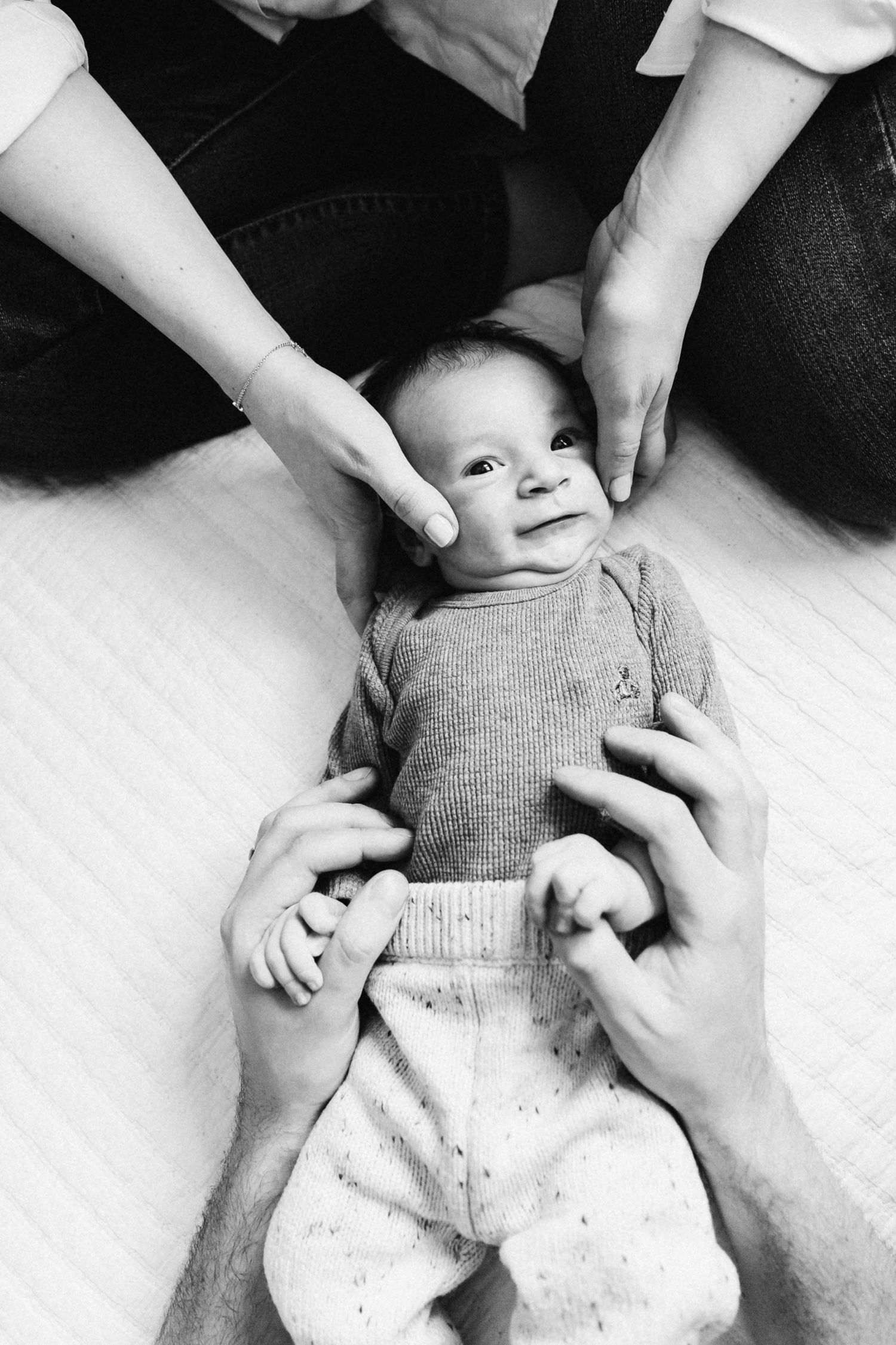 A black and white portrait of a smiling baby lying on their back being held by multiple hands.