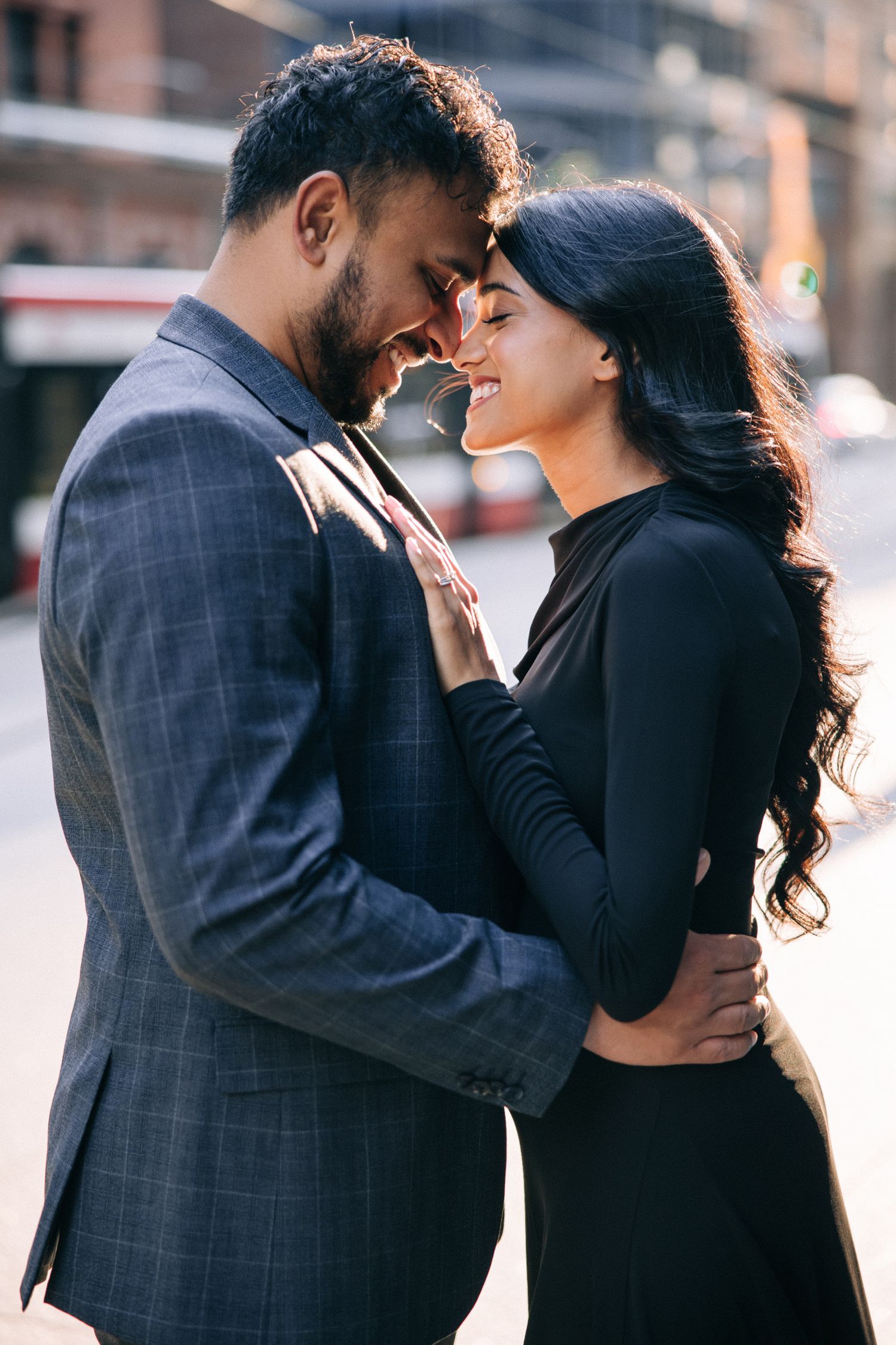 A romantic couple in formal attire share an intimate moment on a city street in warm sunlight.