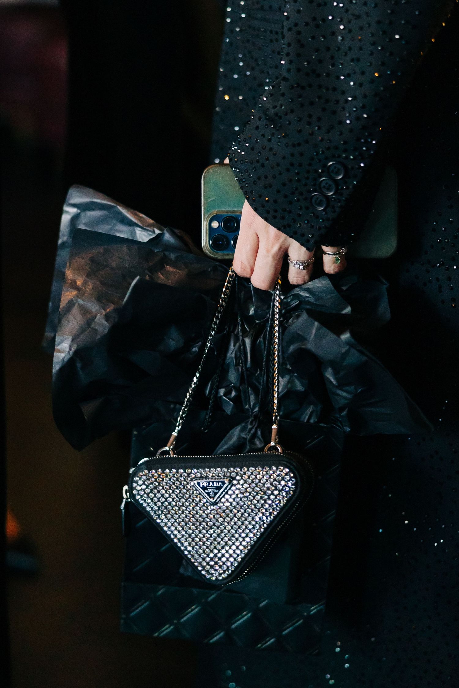 Hand holding a small triangle-shaped crystal-embellished Prada handbag with chain strap against dark background.