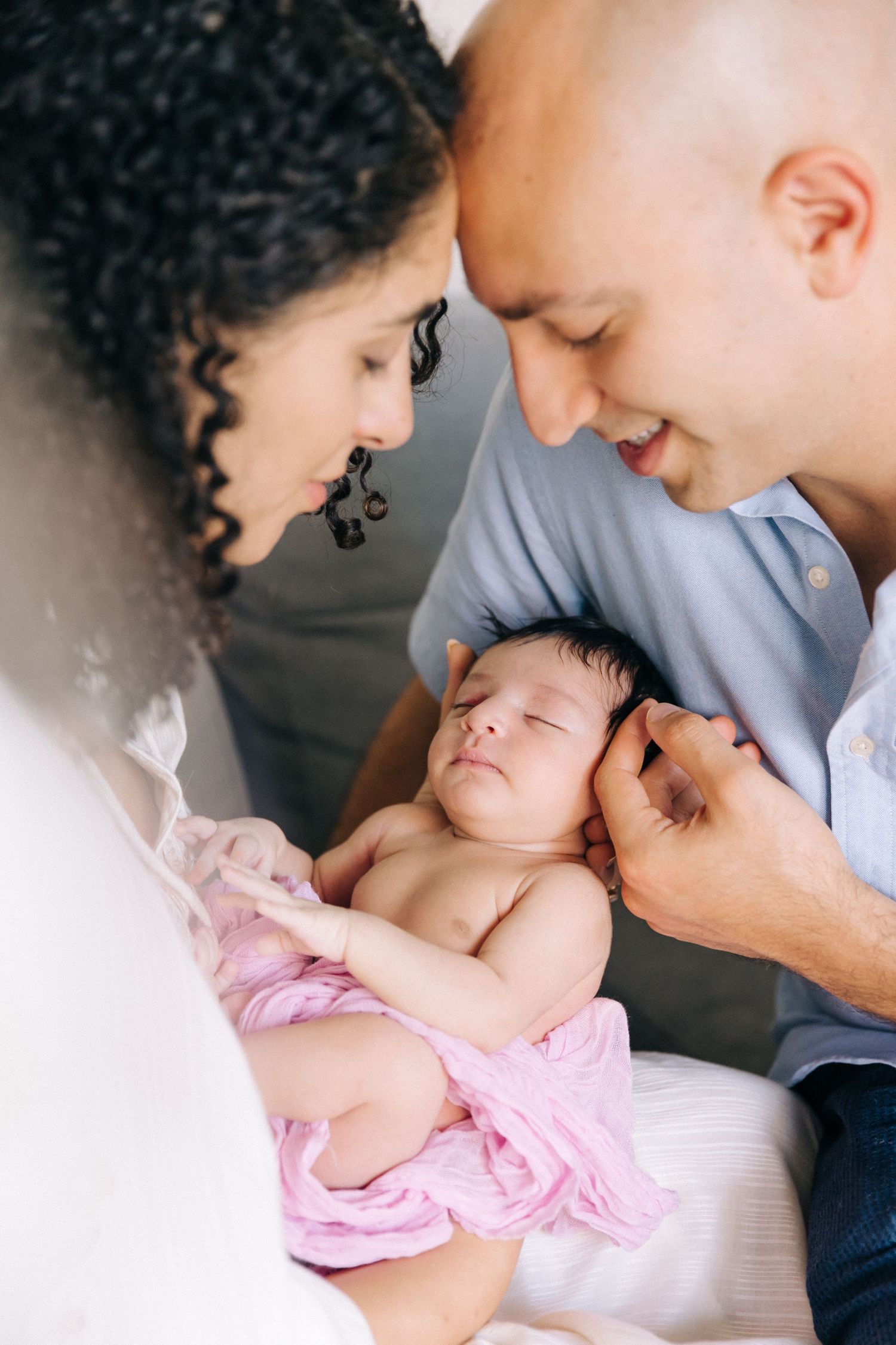 Parents lovingly gaze at their newborn baby swaddled in pink blanket during intimate family moment.