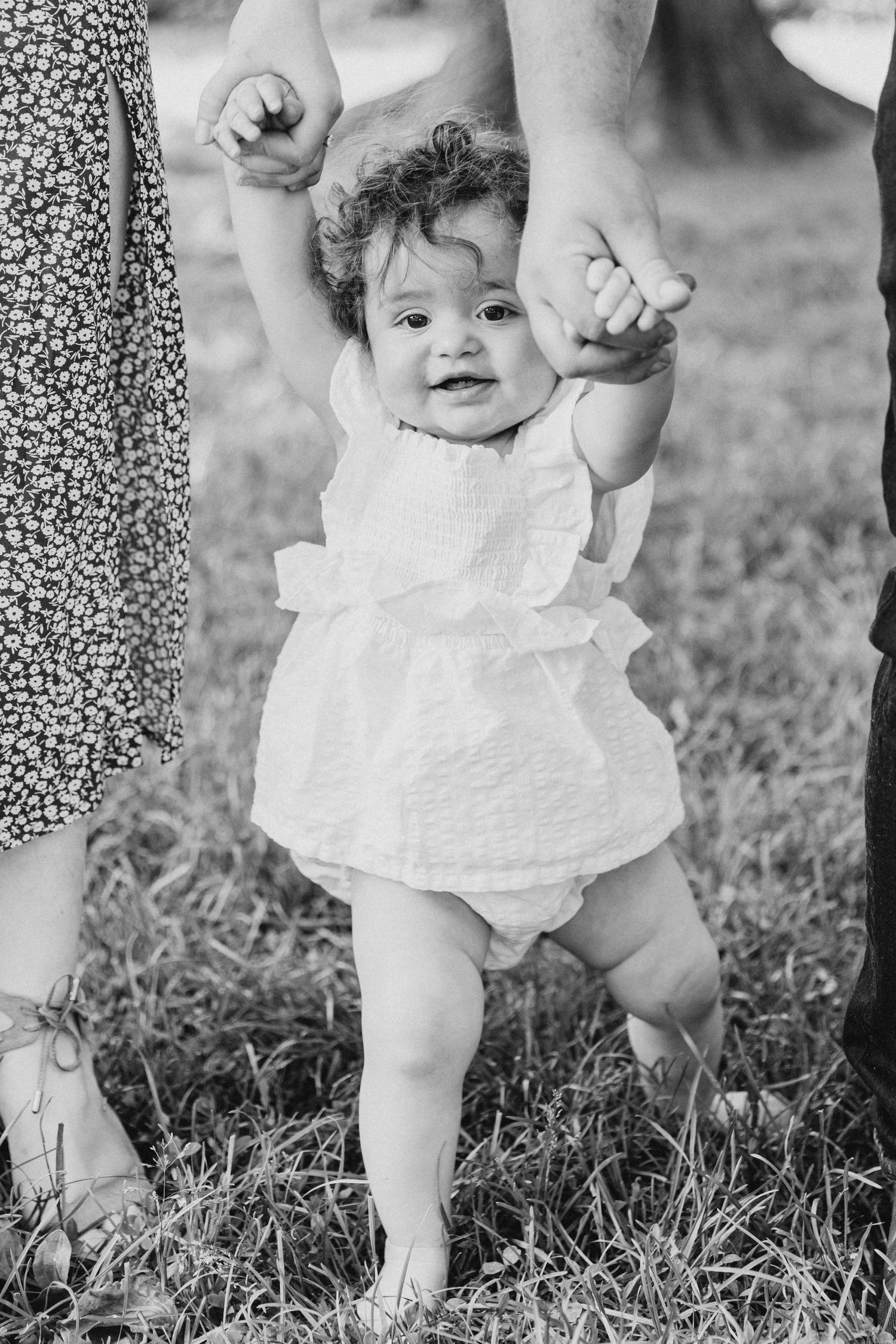 Black and white photo of a smiling baby taking first steps while holding hands.