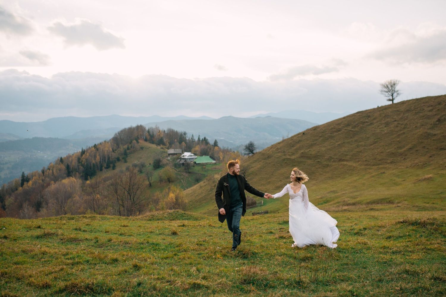 A couple in wedding attire runs together across a scenic mountain meadow with rolling hills and cloudy skies in the background.