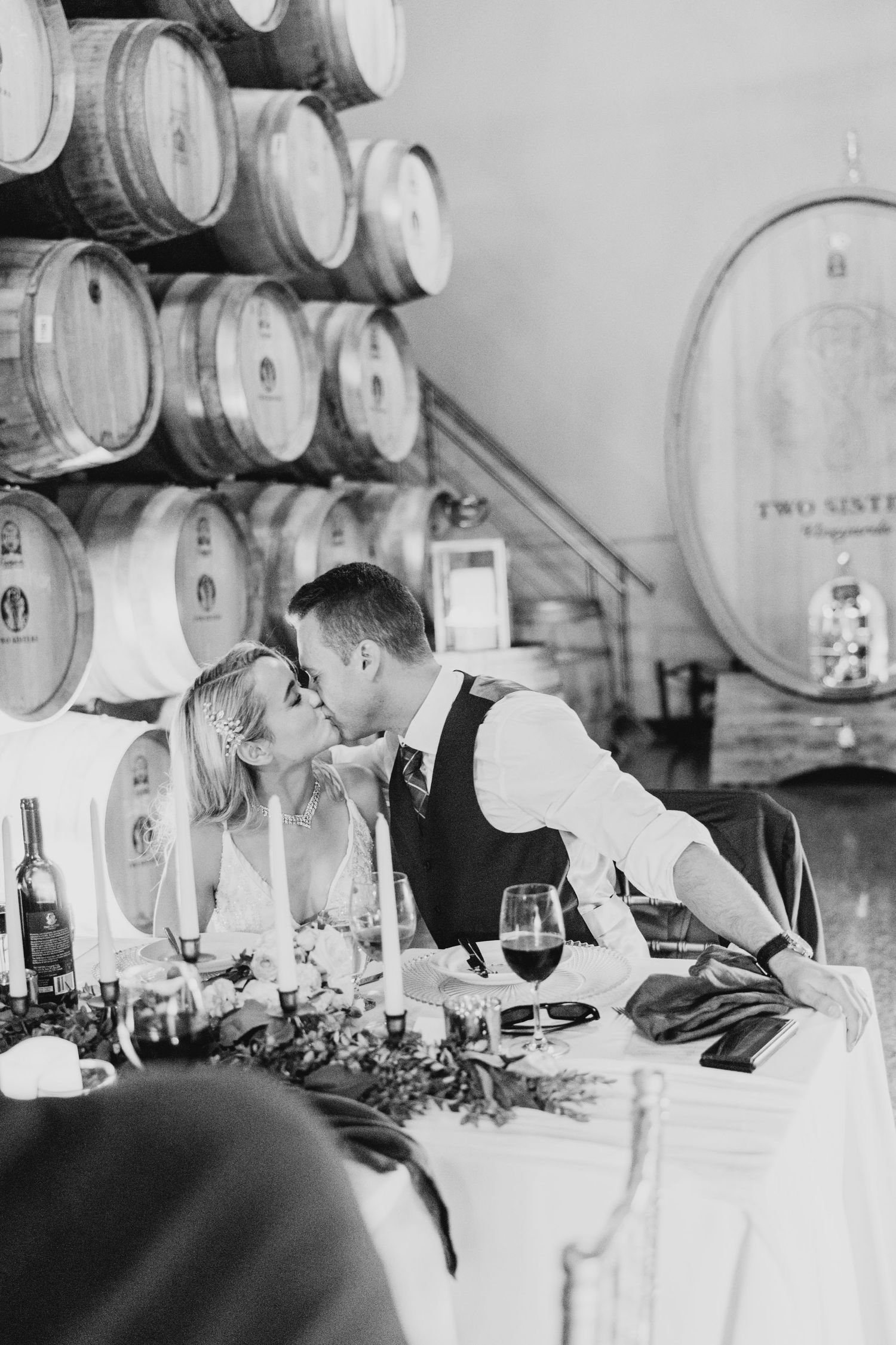 A romantic black and white photo of a couple sharing an intimate moment at a wine barrel-lined venue during a celebration.