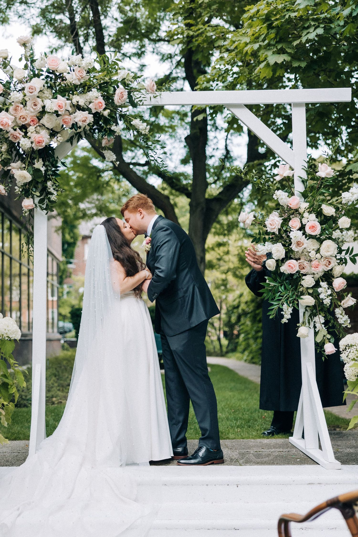 A romantic wedding ceremony under a white arbor decorated with pink and white roses in a lush garden setting.