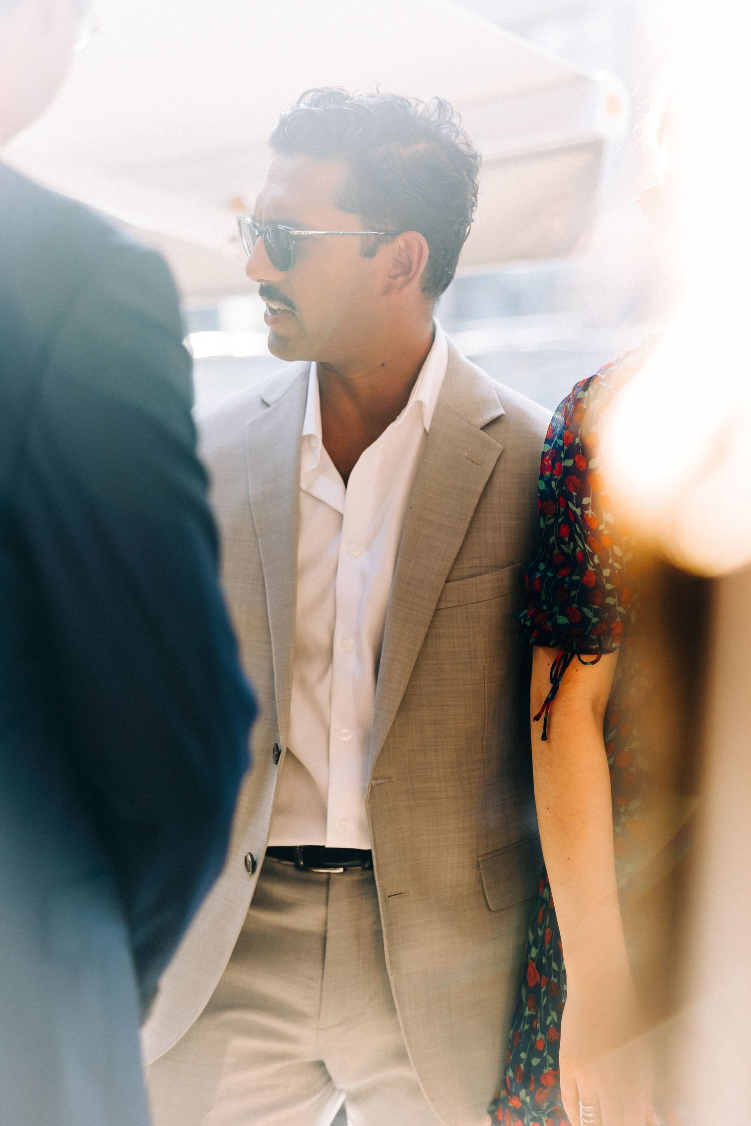 A person in a beige suit and sunglasses stands among others in a sun-drenched setting.