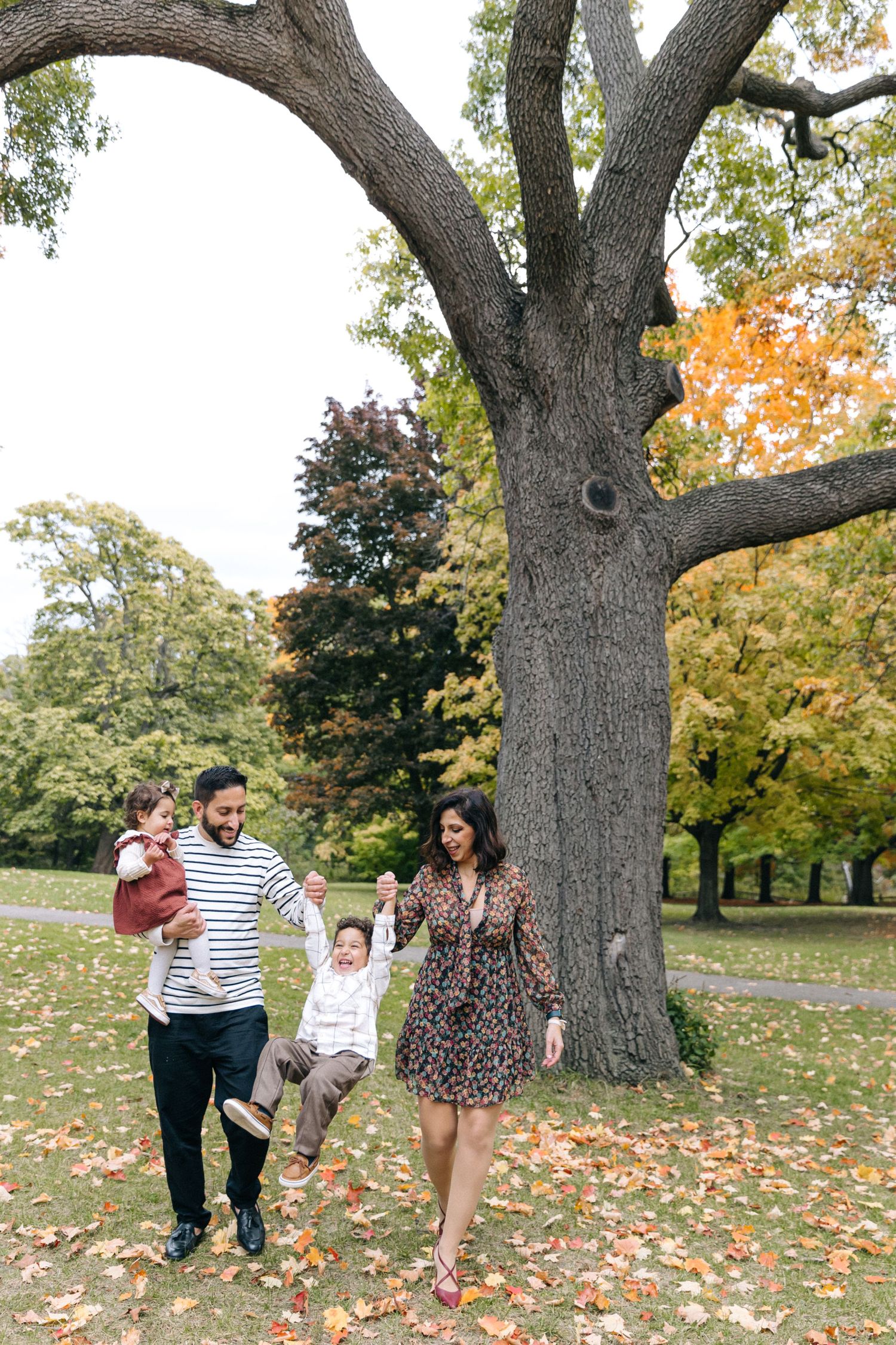 A family walks together under a large autumn tree with fallen leaves on the ground.