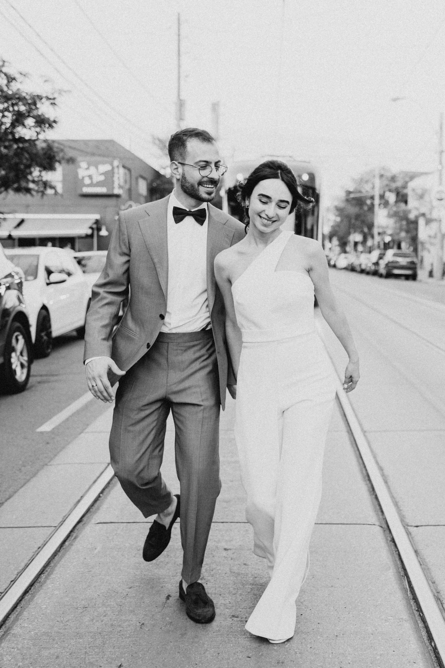 A stylish couple in formal attire walks down a city sidewalk with train tracks and parked cars visible in black and white.