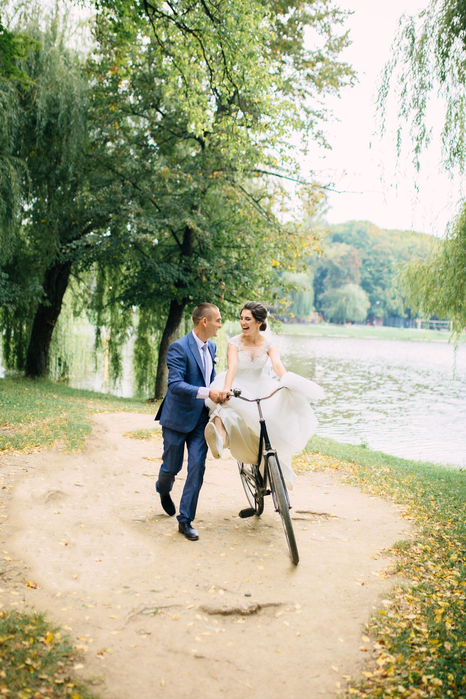 Couple in wedding attire walks together with a bicycle along a scenic lakeside path under weeping willow trees.