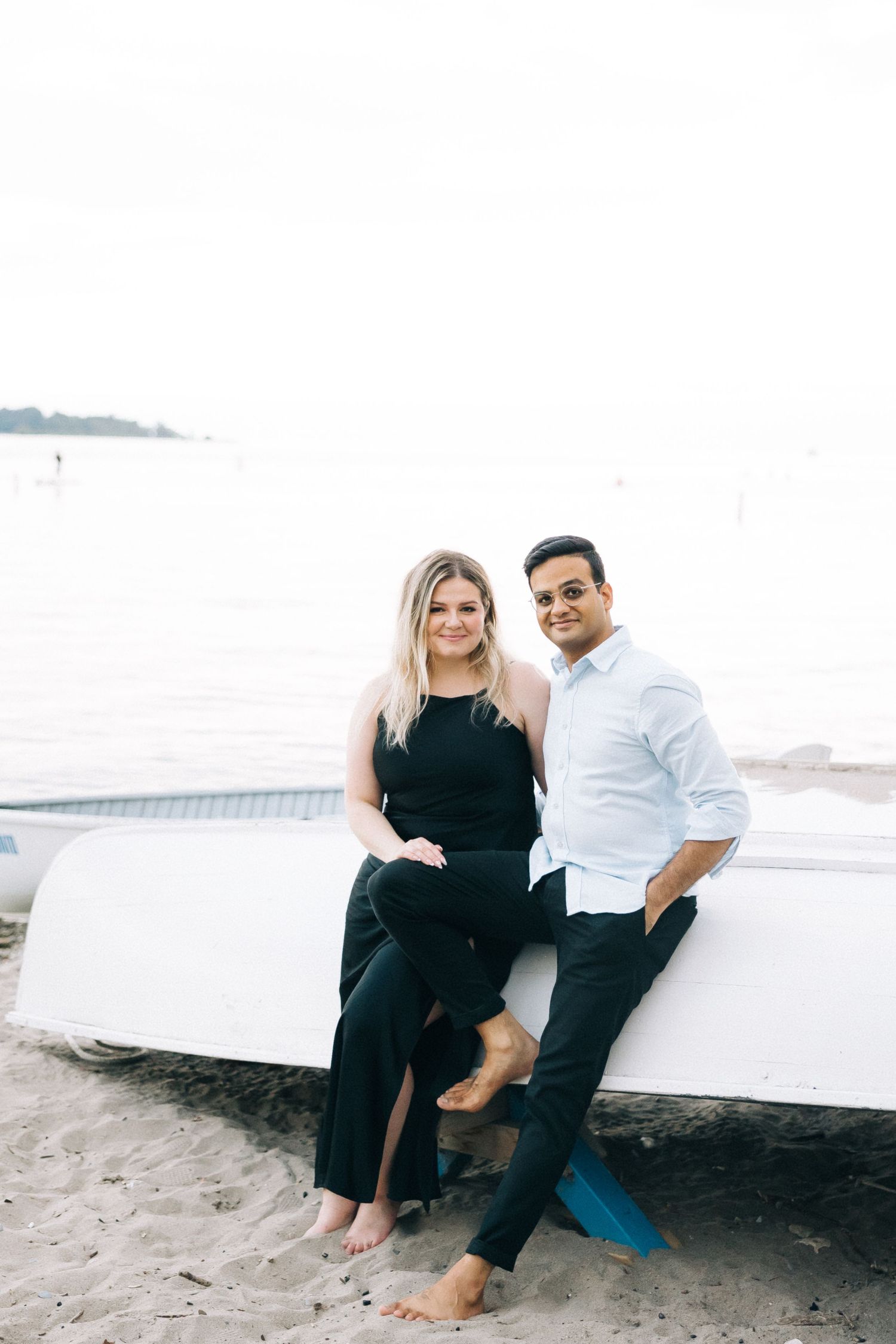 A romantic couple poses together against a boat on a sandy beach.