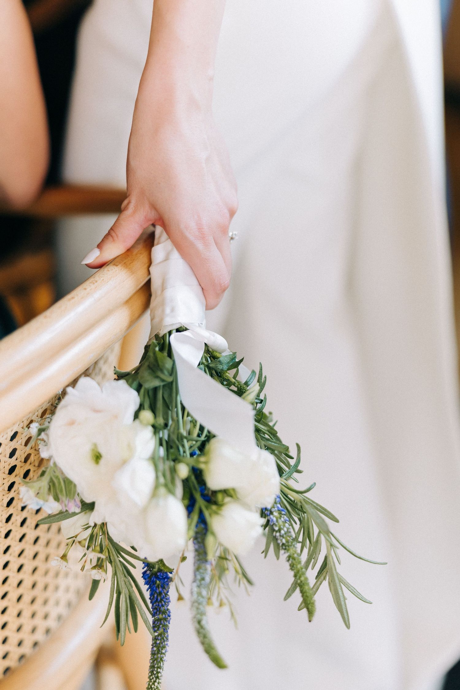 A delicate white bouquet with rosemary sprigs is held against a rattan chair, with a glimpse of a flowing white dress.