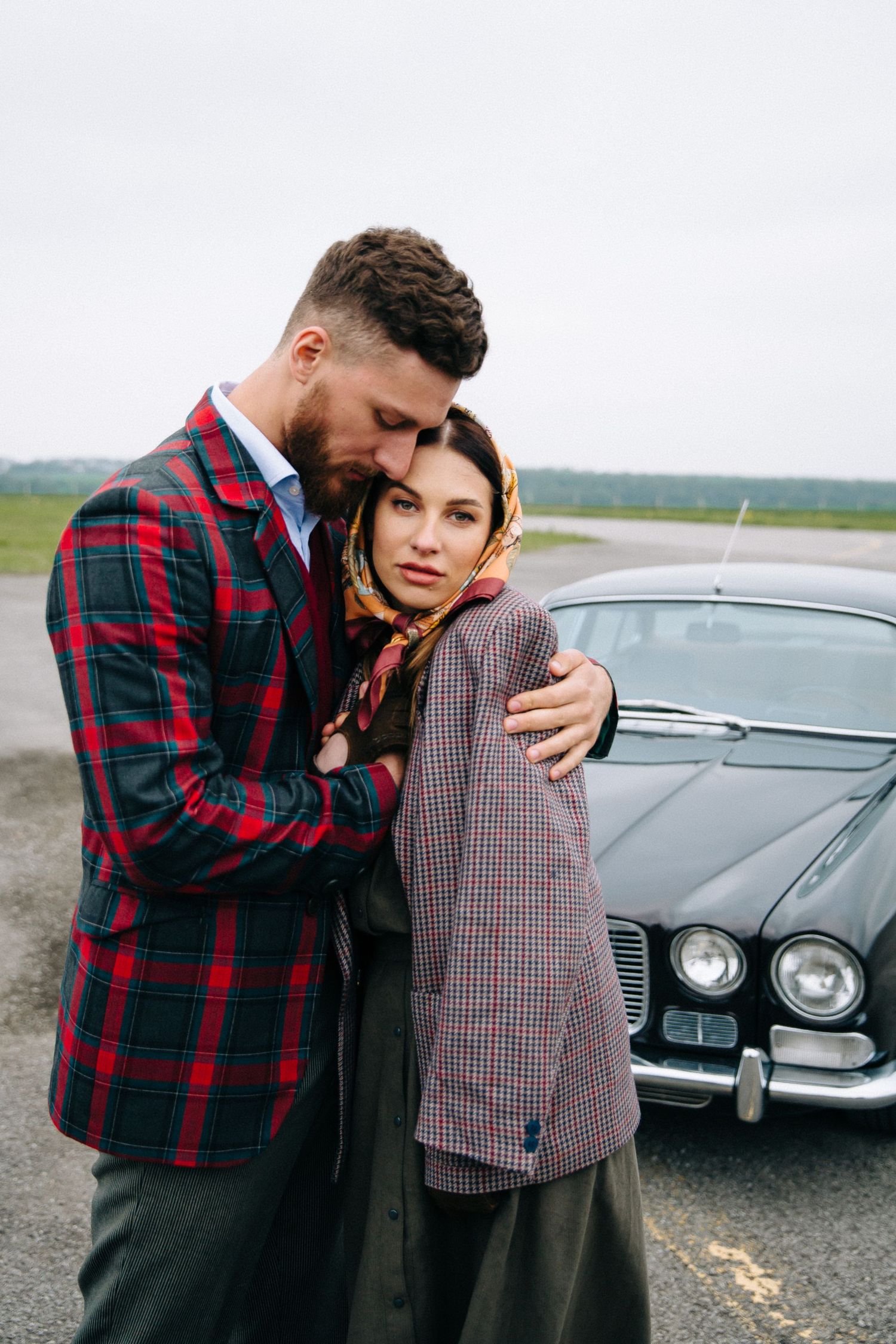 A couple in vintage-inspired outfits embraces near a classic car.