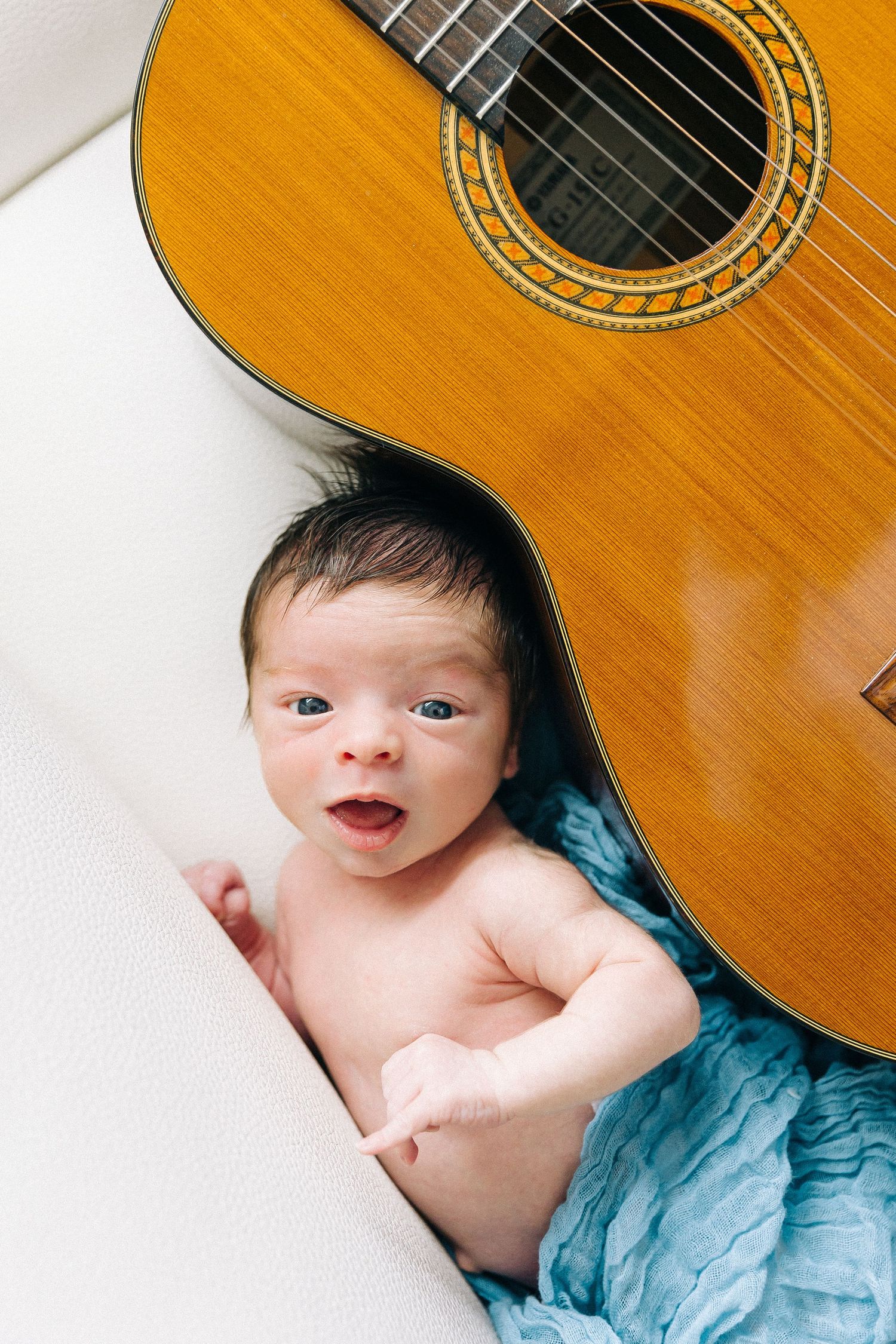 A creative portrait of a baby lying next to an acoustic guitar on white fabric.