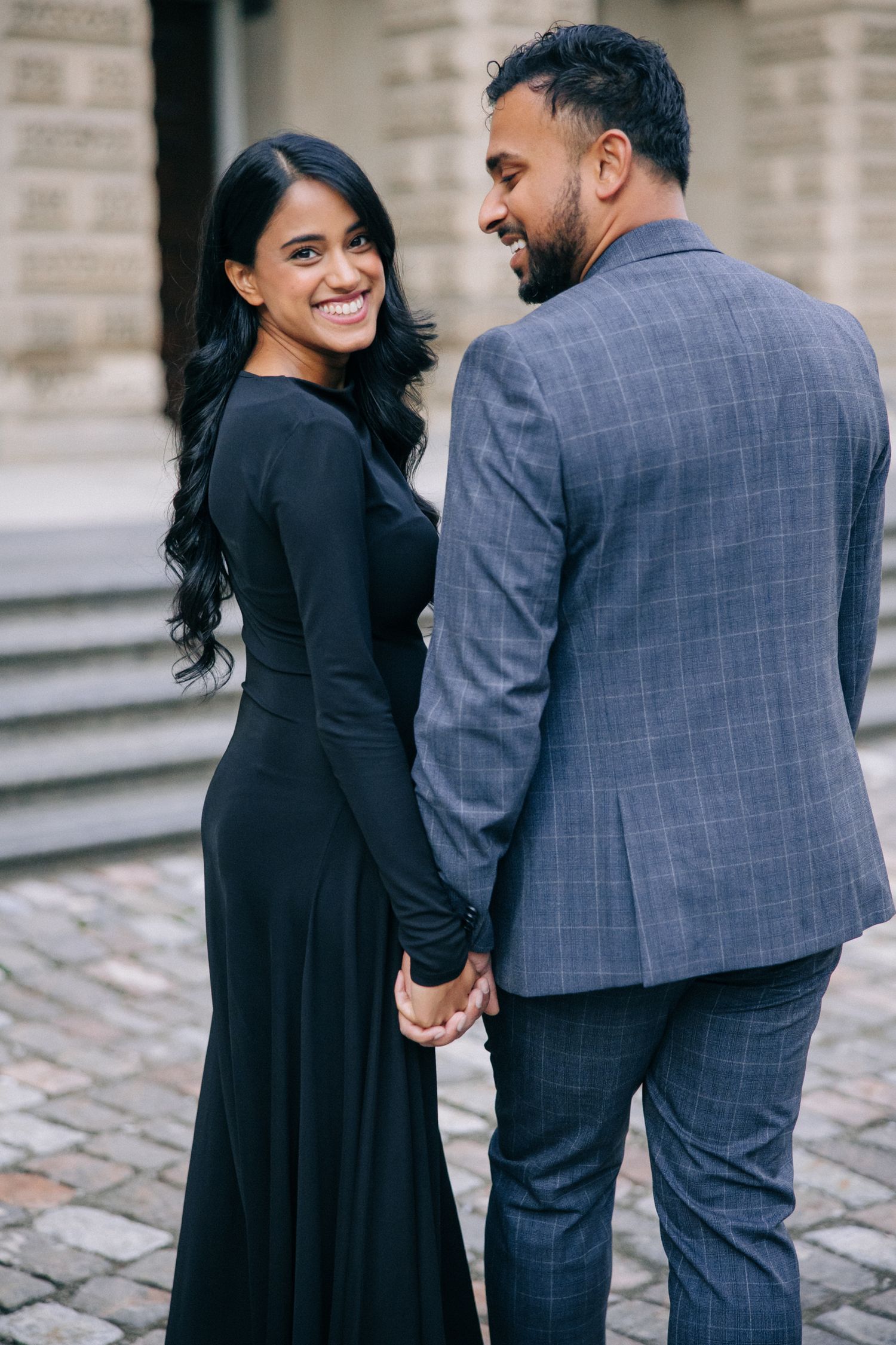 A couple holds hands while sharing a romantic moment outdoors, dressed elegantly in black and navy formal attire.