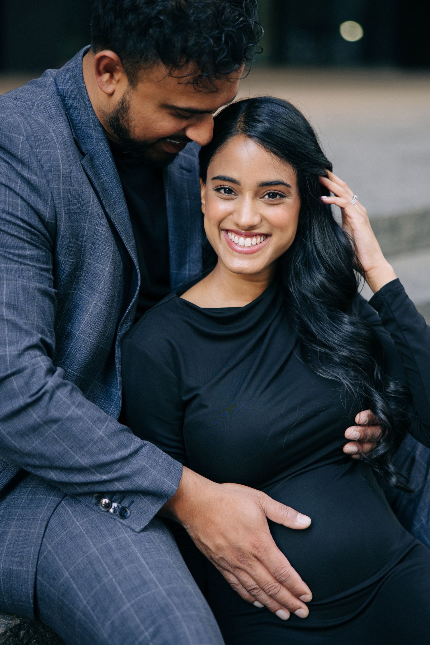 A couple shares an intimate moment during a maternity photo session, with her wearing black and him in a gray plaid suit.